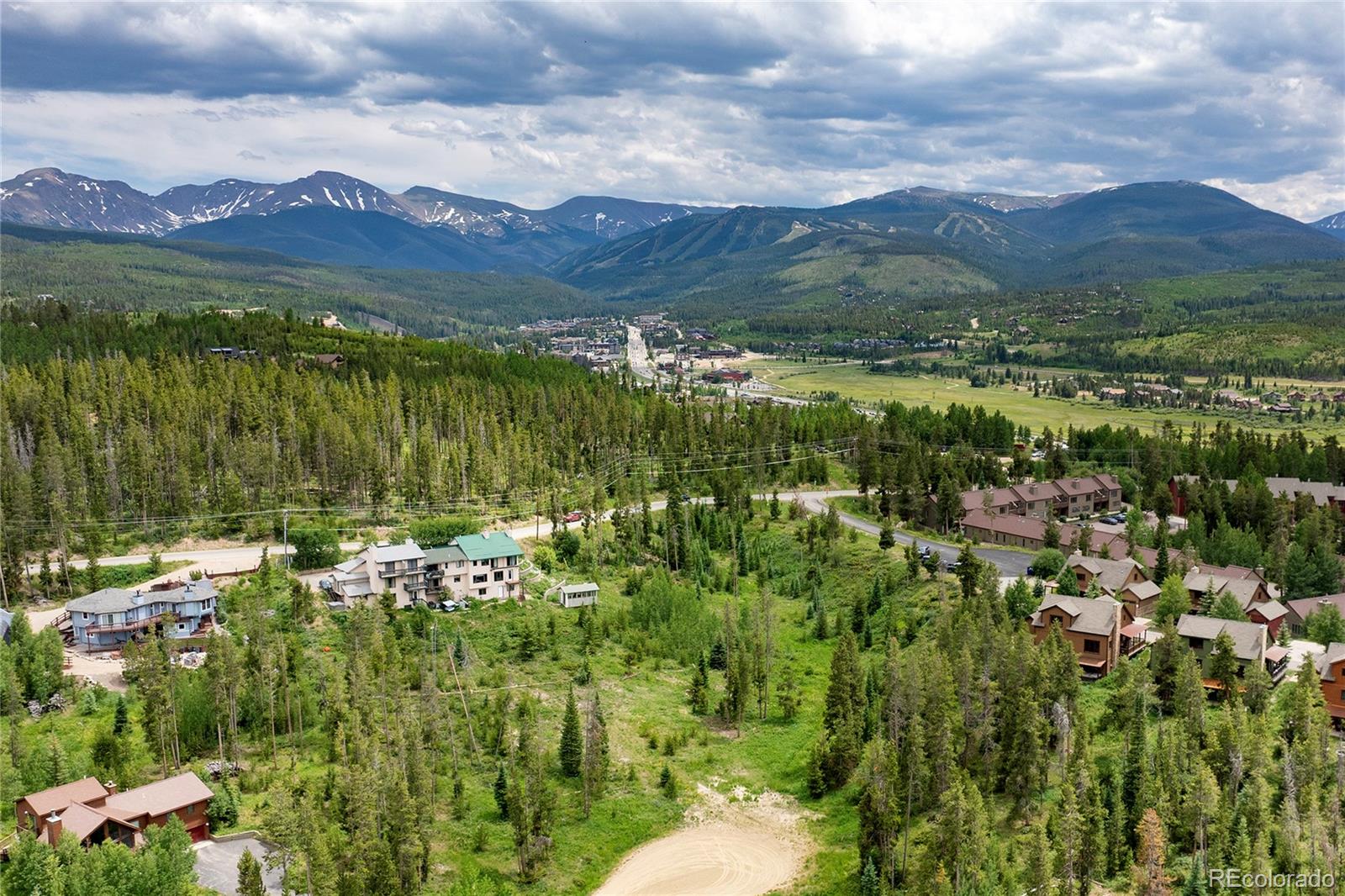 Aerial view of a mountain valley with homes, forest, a distant town, and snow-patched peaks.