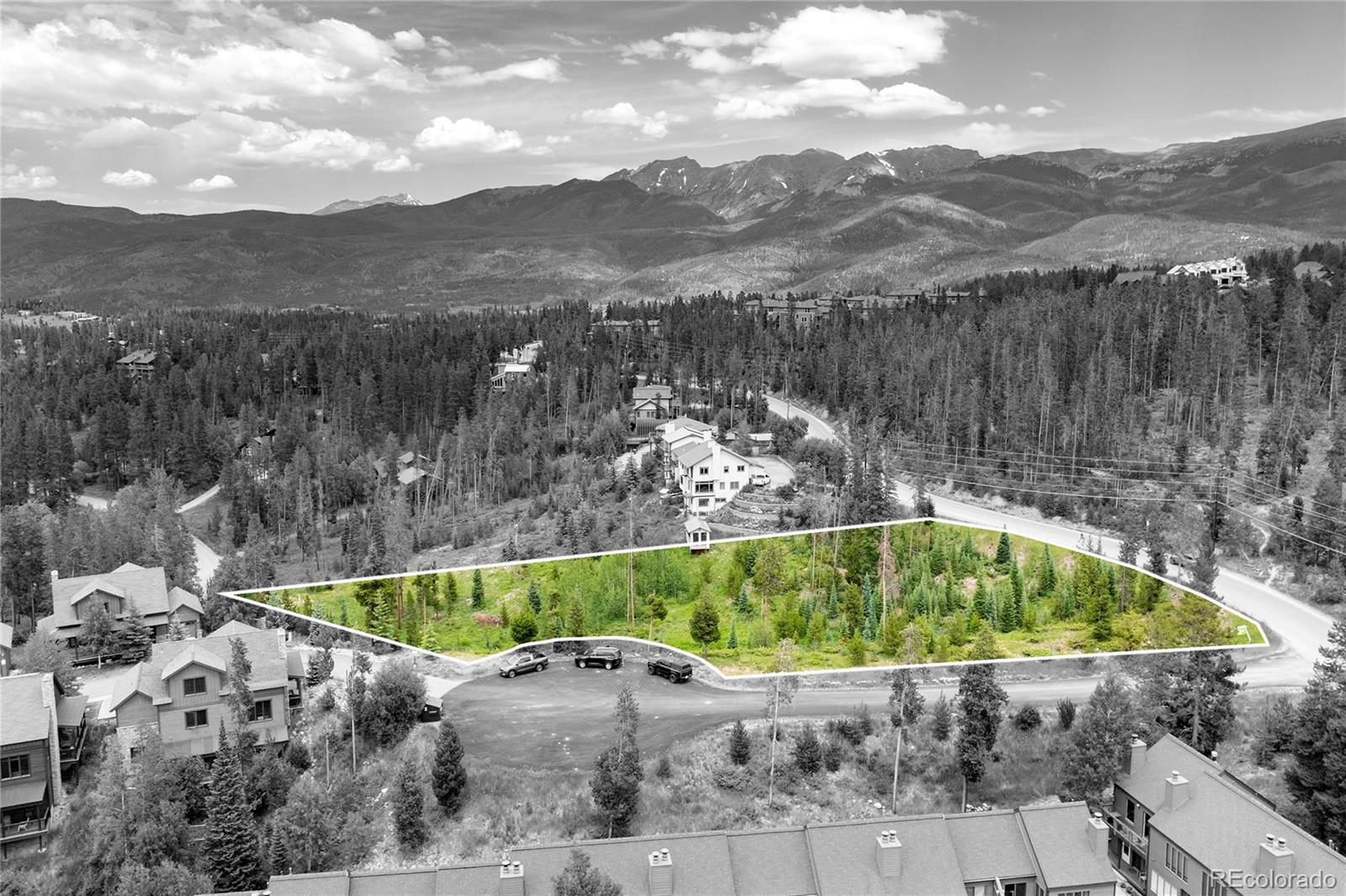 Aerial view of a wooded vacant lot outlined in white, with nearby homes and mountain views.