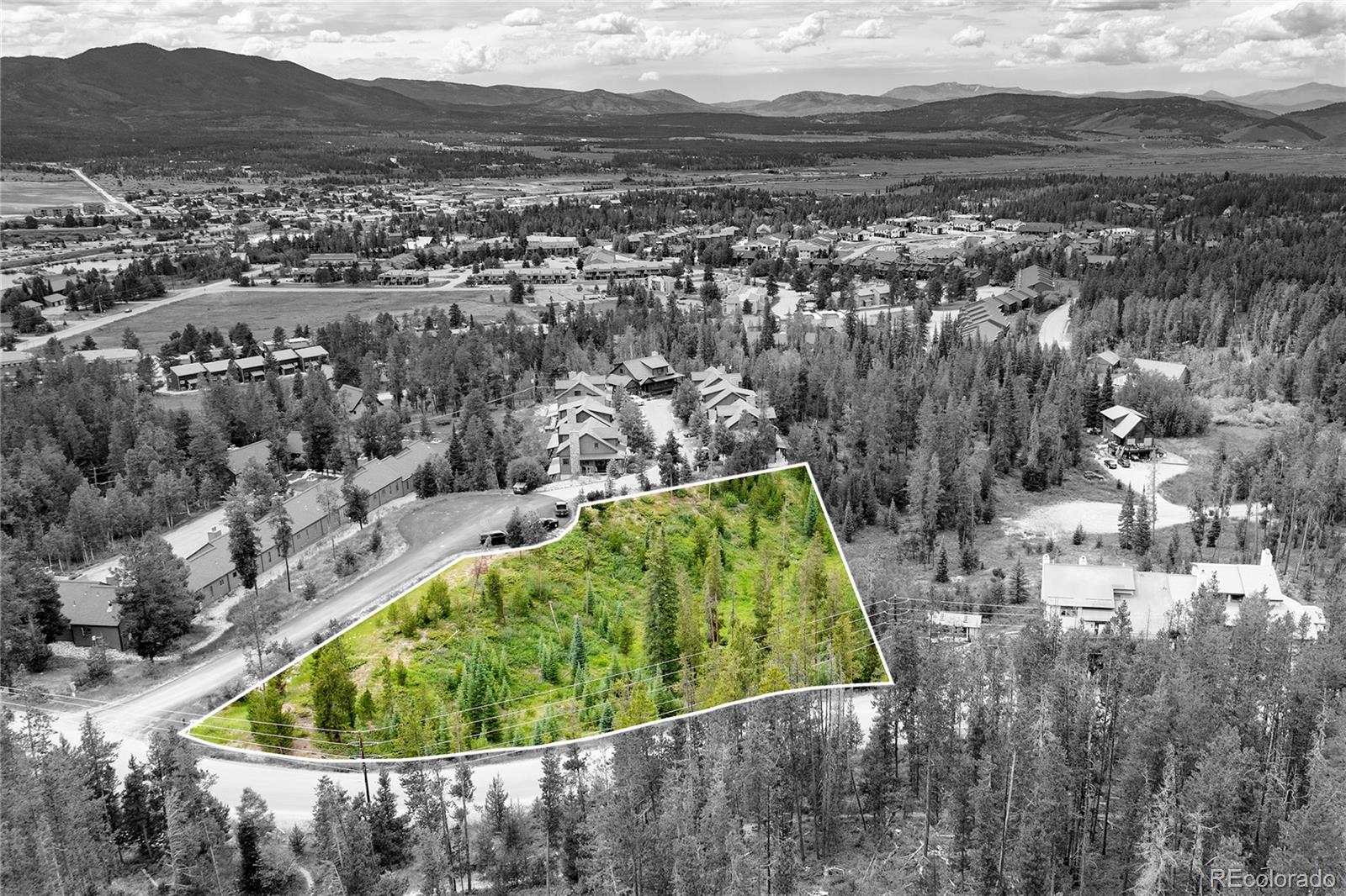 Aerial view of a wooded parcel outlined in white, with surrounding homes and distant mountains.