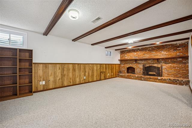 Empty basement living room with wood-paneled walls, exposed ceiling beams, and a brick fireplace wall.