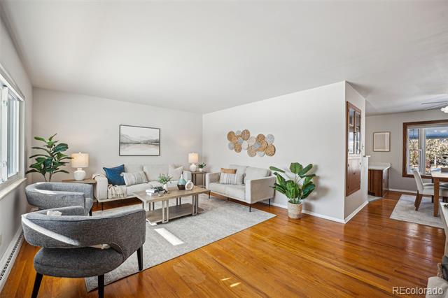 Staged living room with hardwood floors, gray seating, large windows, and an open dining area.