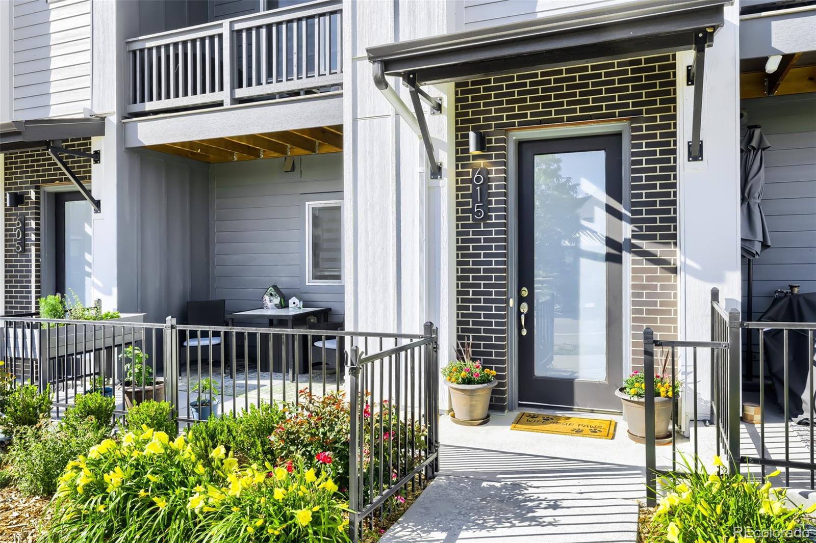 Townhouse entrance with a glass door, covered awning, fenced walkway, and flowering garden.