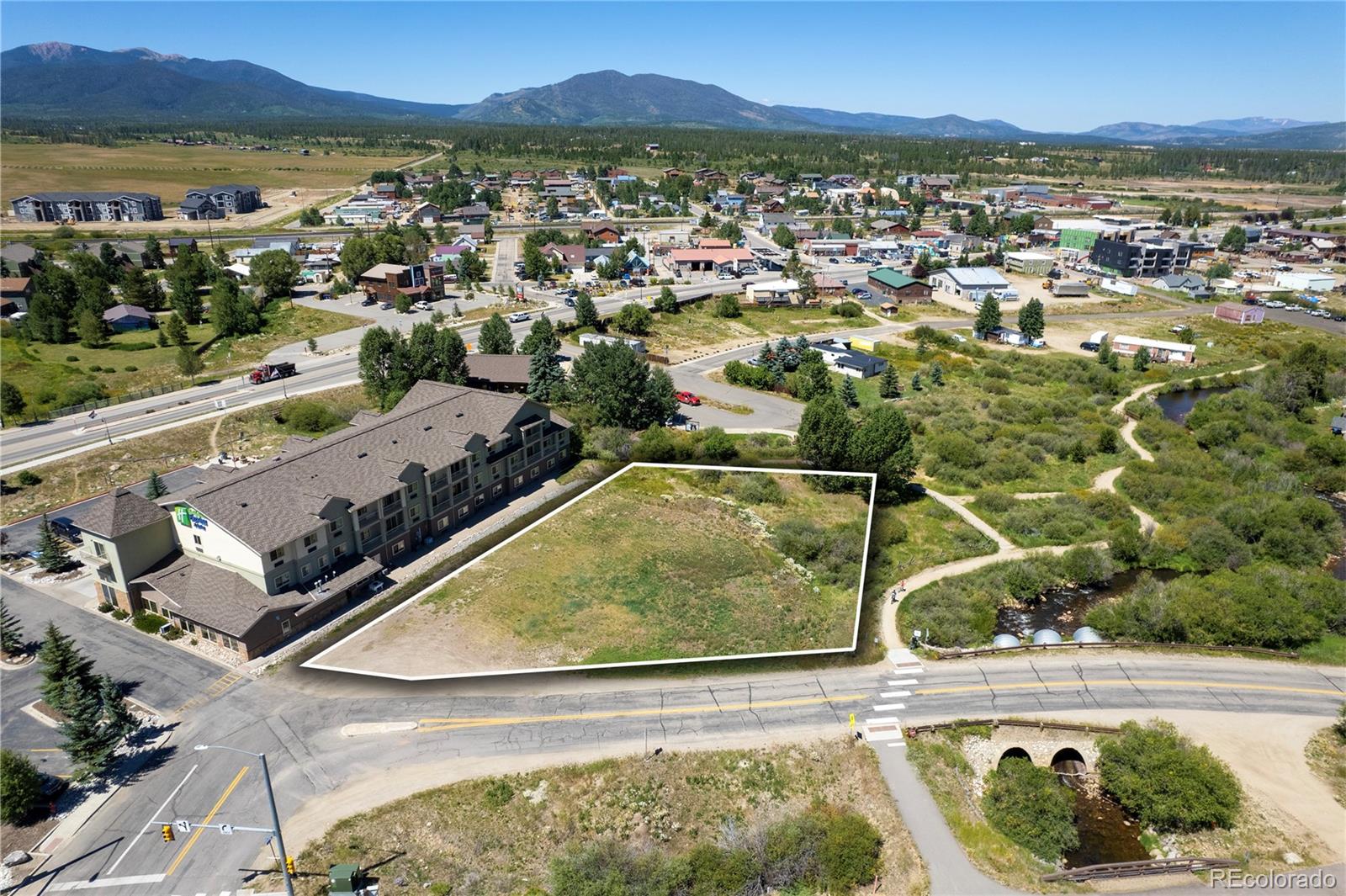 Aerial view of a vacant outlined lot beside a building, with a creek and mountains nearby.