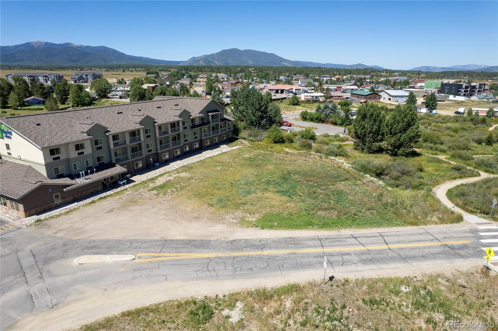 Aerial view of a condominium building, open lot, and Fraser with mountain ranges beyond.