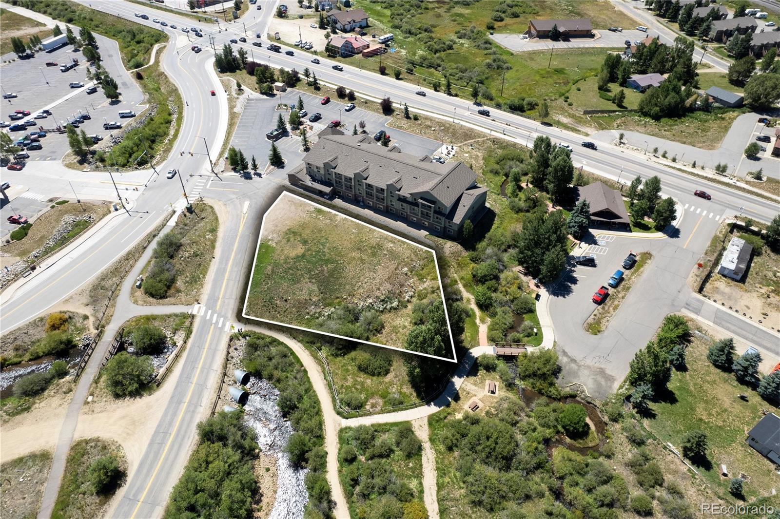 Aerial view of an outlined vacant parcel beside a creek, roads, and a multistory building.