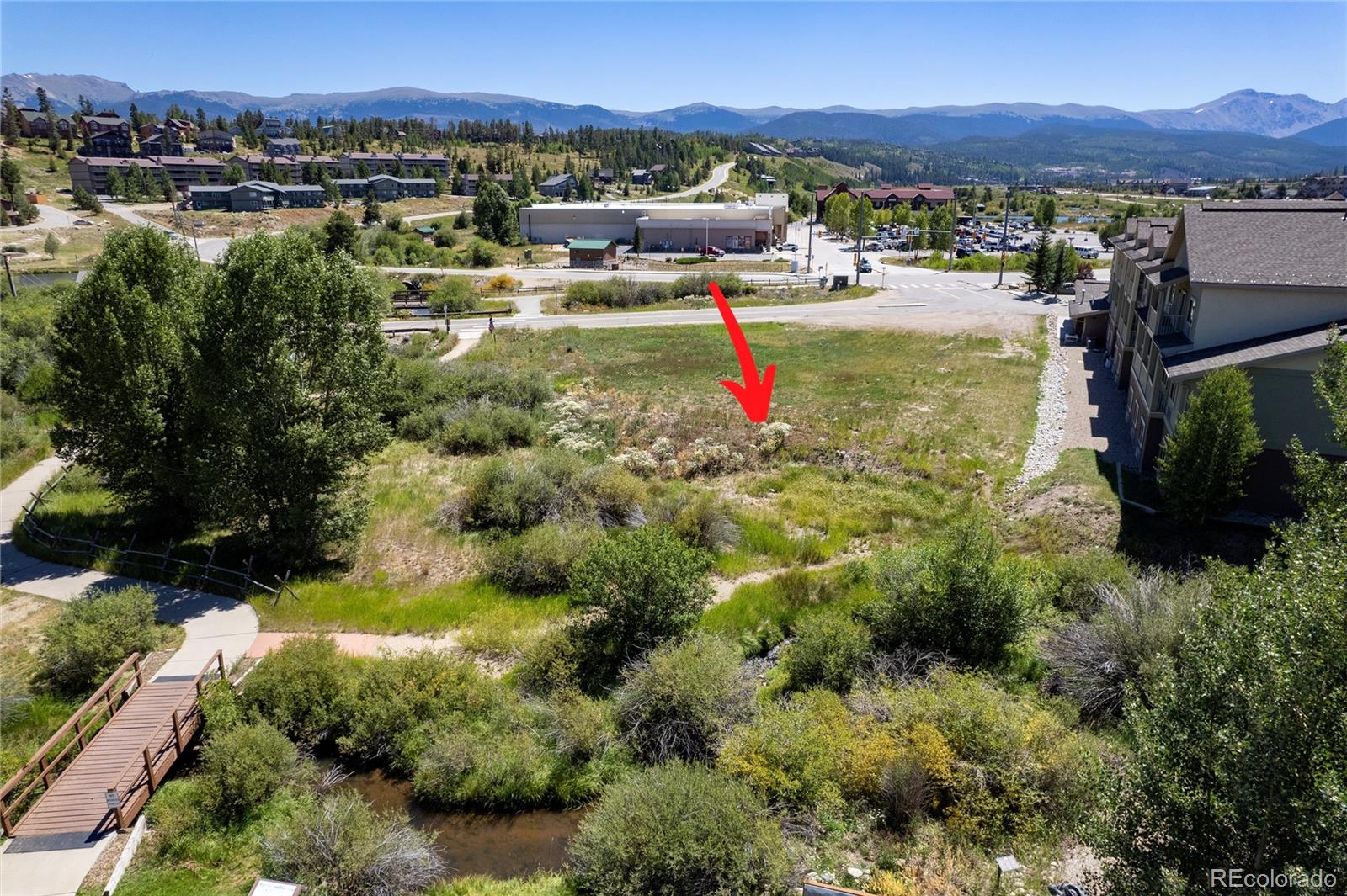 Vacant lot marked by a red arrow, with a creek and footbridge in the foreground and mountains beyond.