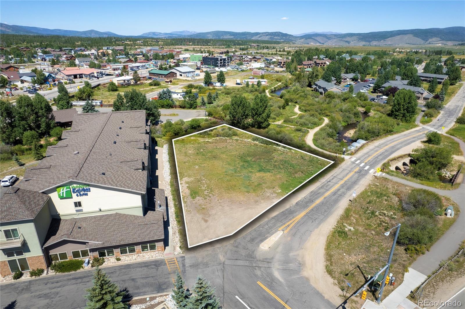Outlined vacant parcel beside a road, with a hotel nearby and mountains in the distance.