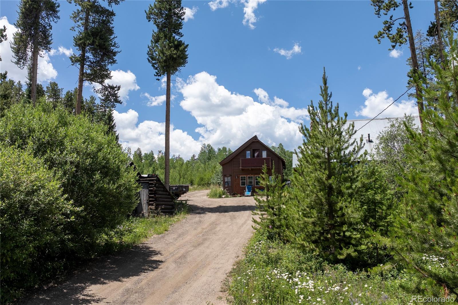 Brown cabin with an upper balcony at the end of a dirt driveway among tall pine trees.