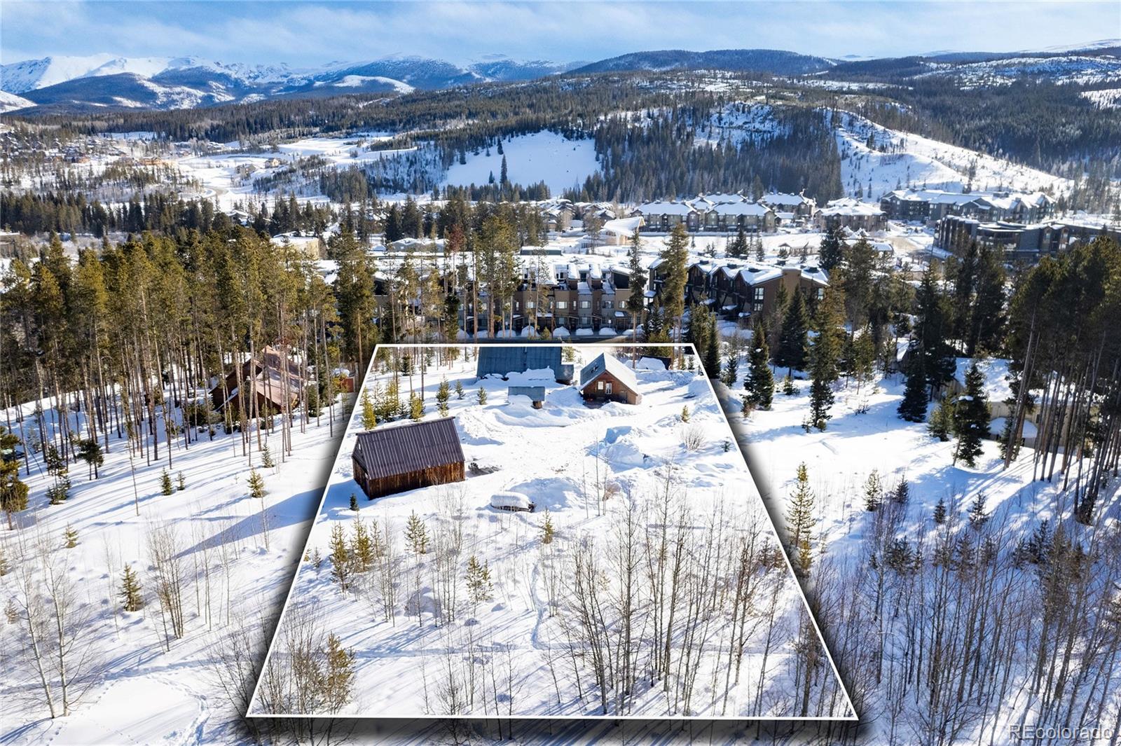 Snow-covered parcel with small buildings, pine trees, and resort buildings beneath forested mountain slopes.