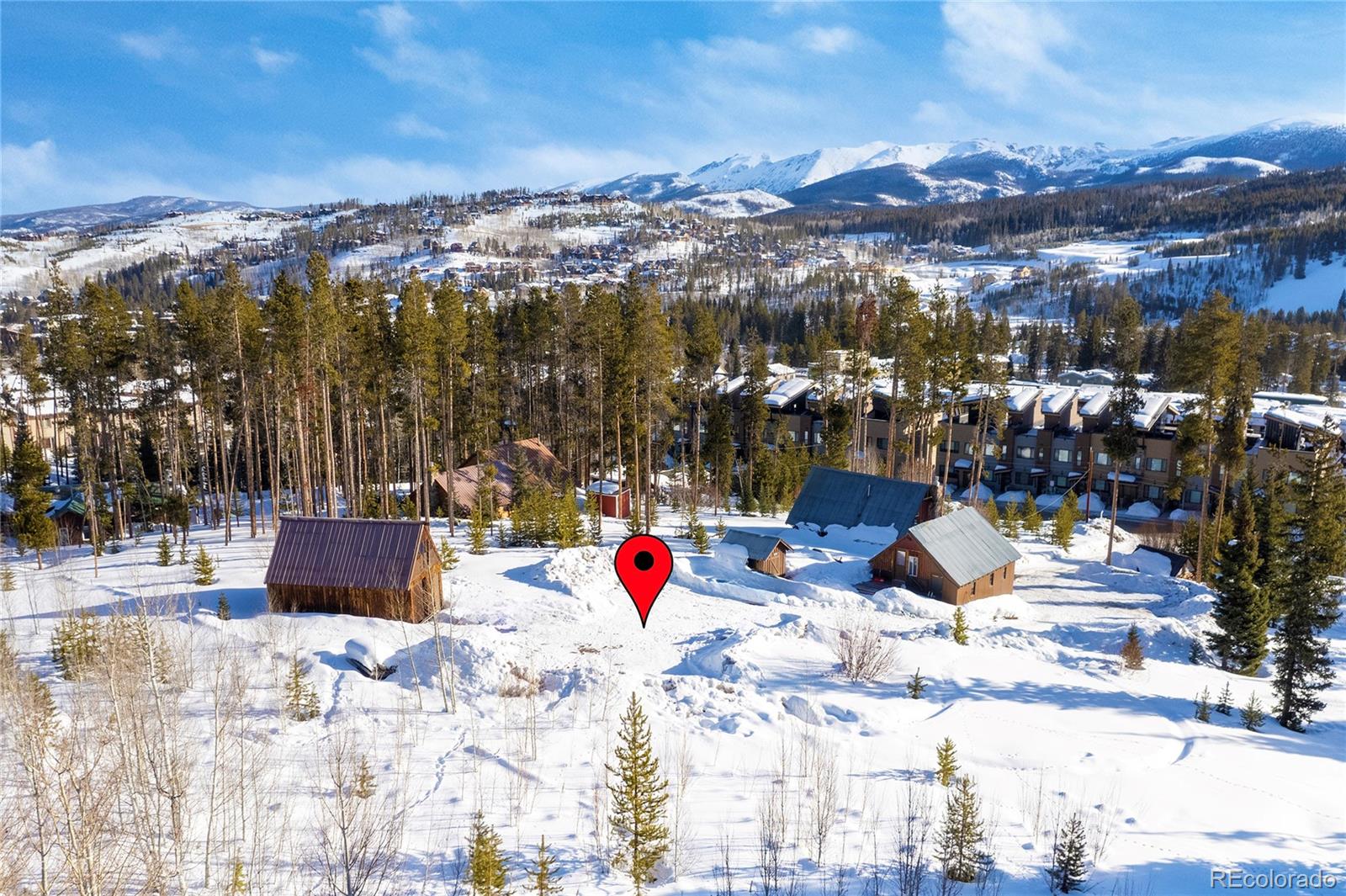 Snow-covered vacant lot marked by a red pin, with evergreen trees, cabins, and mountains beyond.
