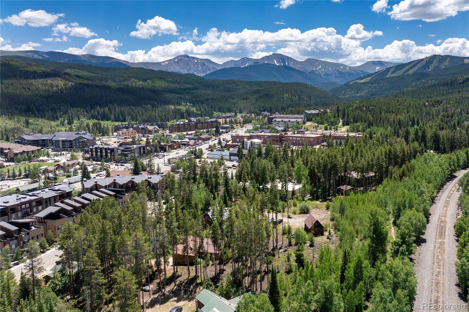 Aerial view of a forested mountain town, with snow-patched peaks and railway tracks.