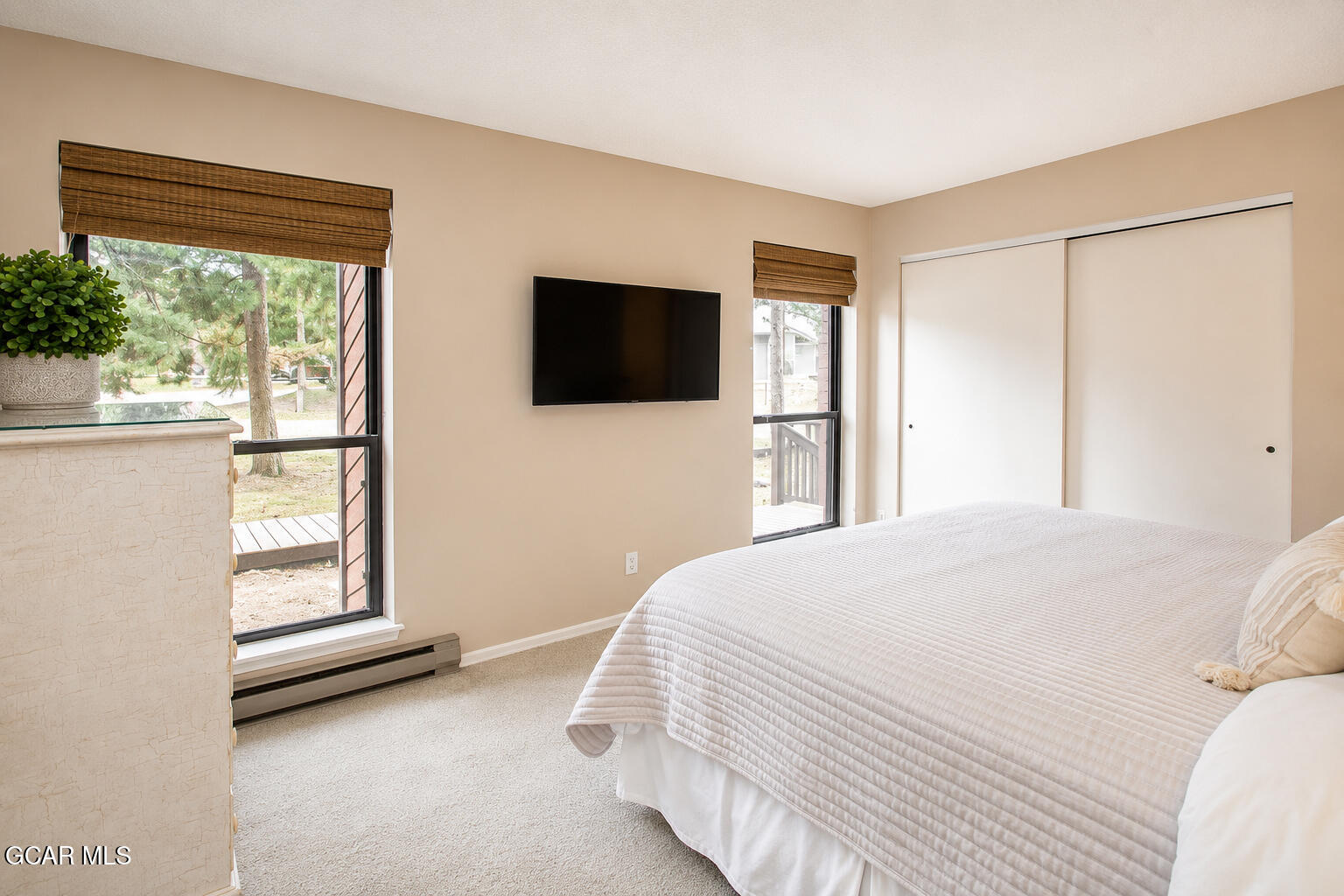 Bedroom with a white quilted bed, wall-mounted TV, and windows overlooking trees.