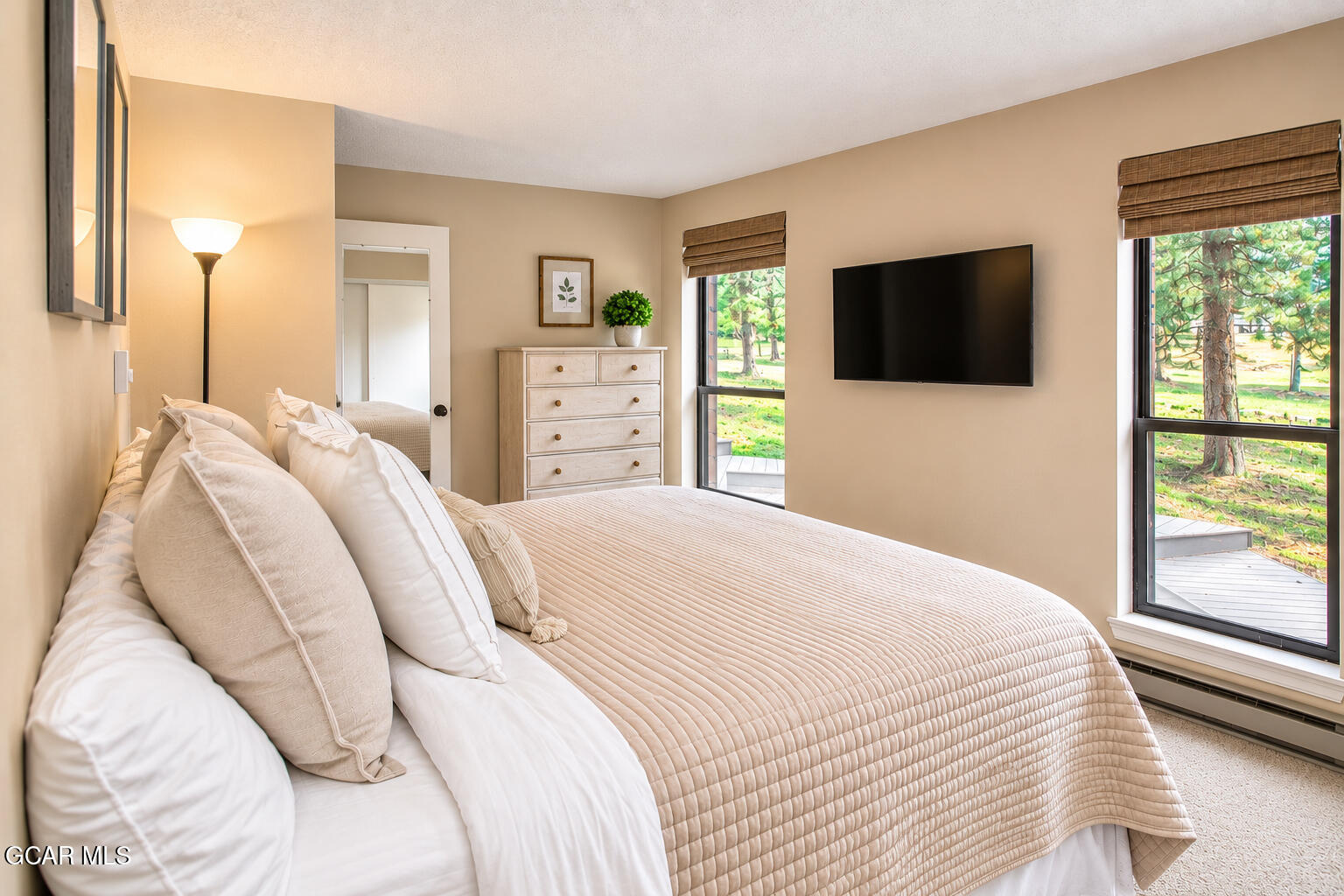 Bedroom with a quilted bed, dresser, wall-mounted TV, and windows overlooking trees.