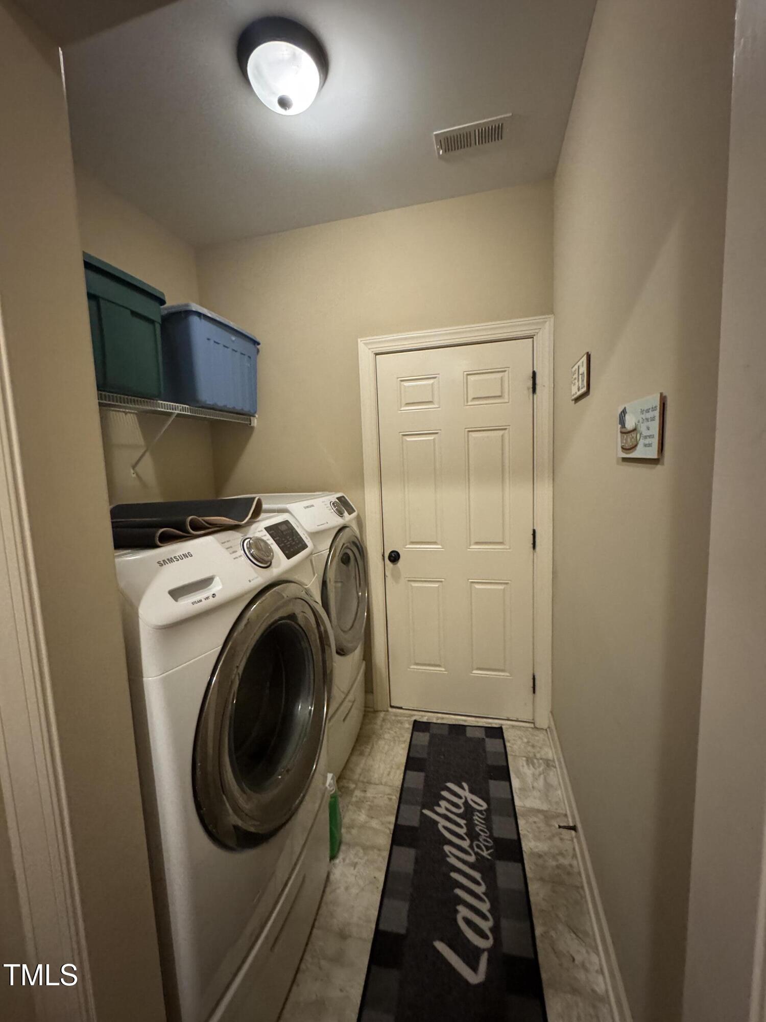 Laundry room with white front-loading washer and dryer, overhead storage shelf, and black rug.