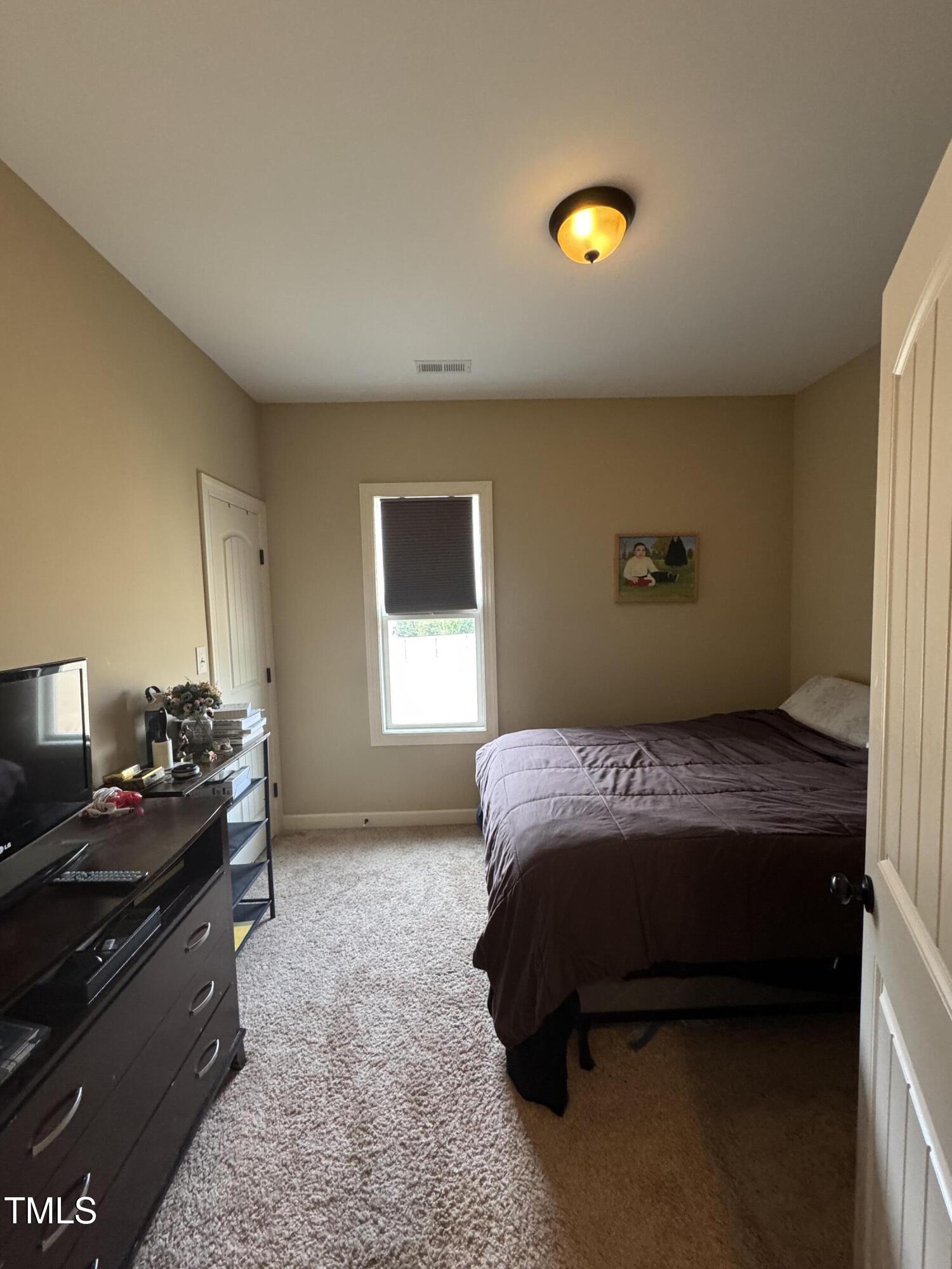 Furnished bedroom with a dark bedspread, carpeted floor, and window with a dark blind.