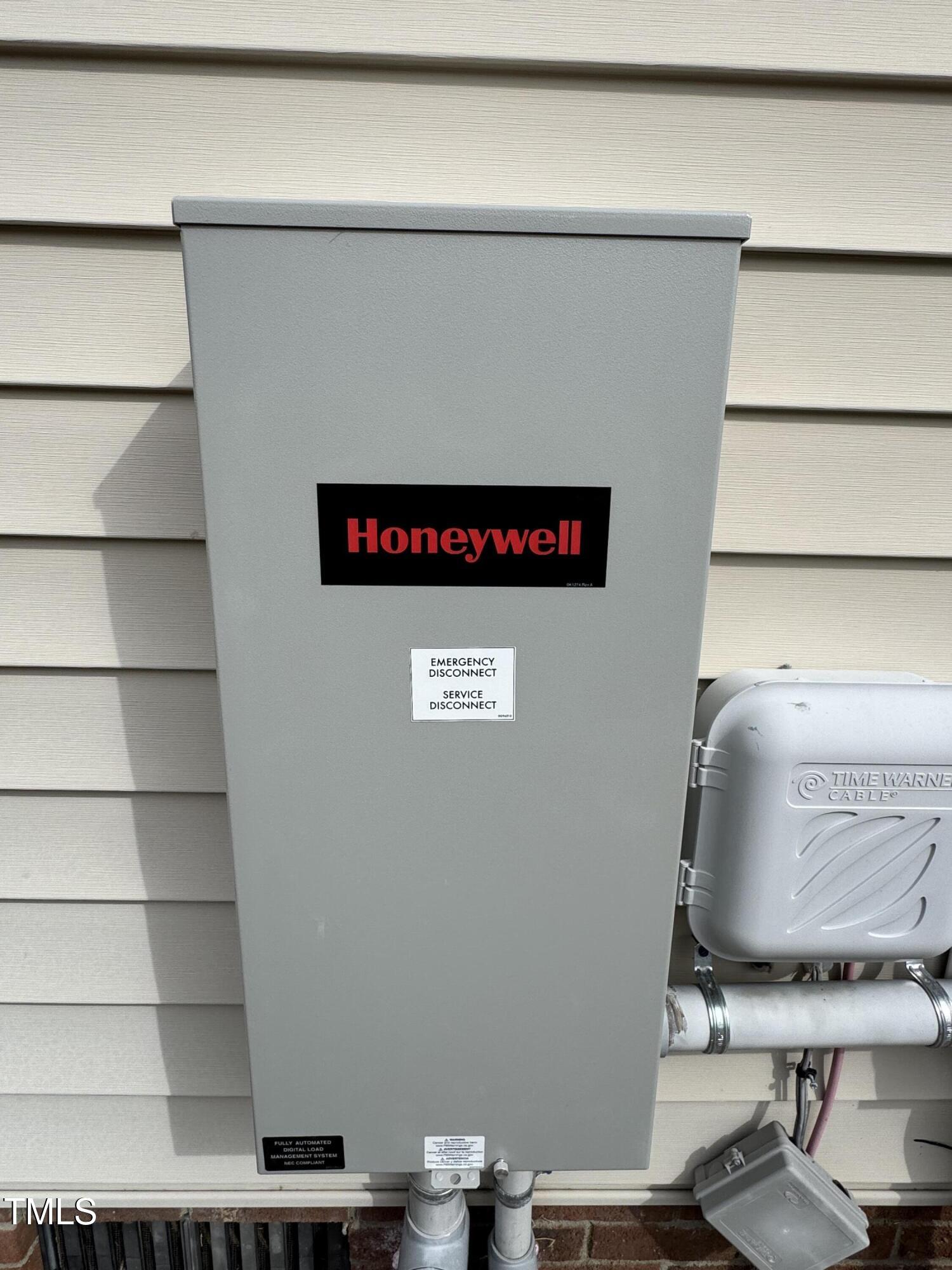 Honeywell electrical disconnect cabinet mounted on house siding beside utility pipes and a cable enclosure.