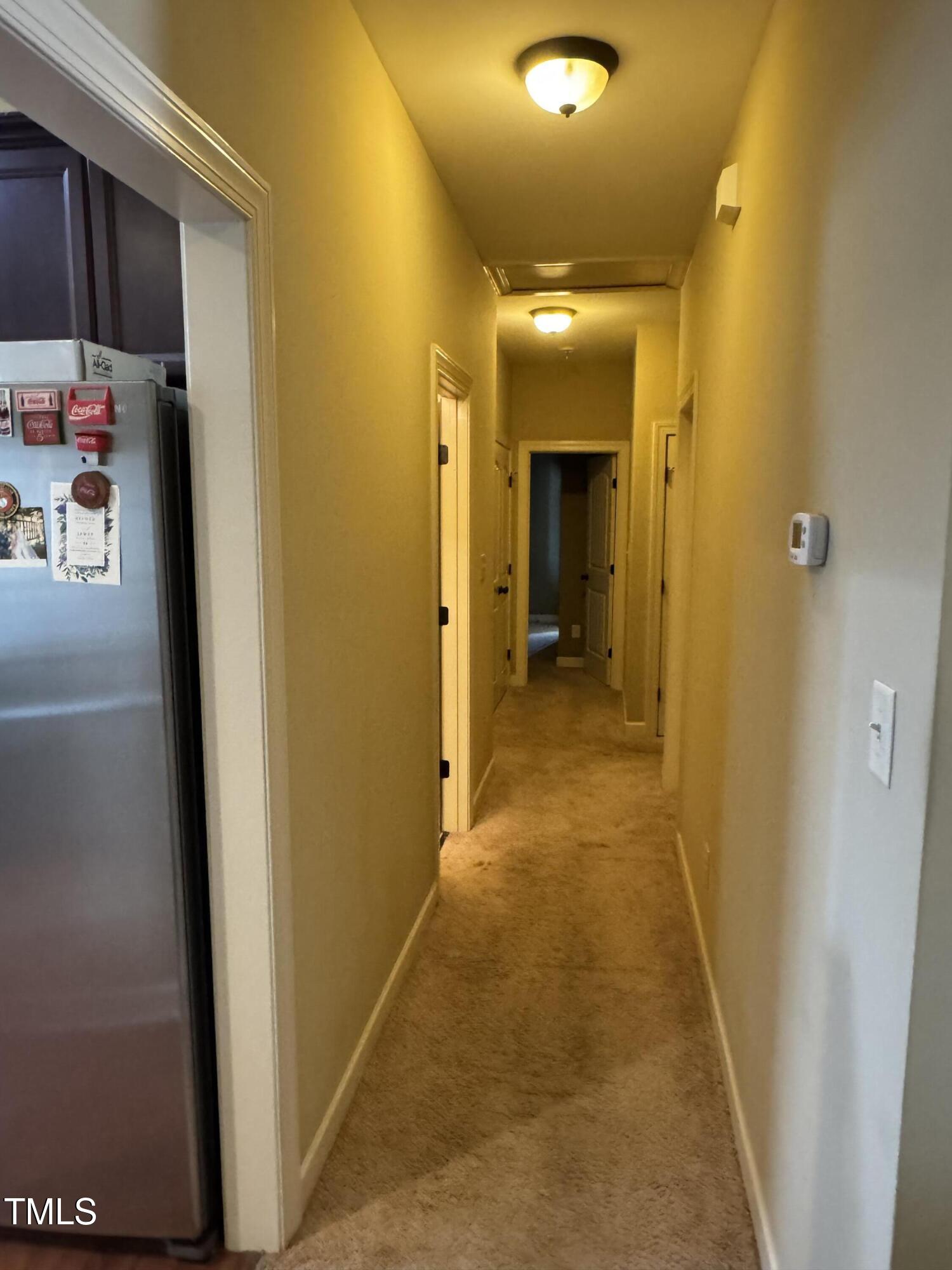 Carpeted hallway with several doorways, ceiling lights, and a refrigerator at the left edge.