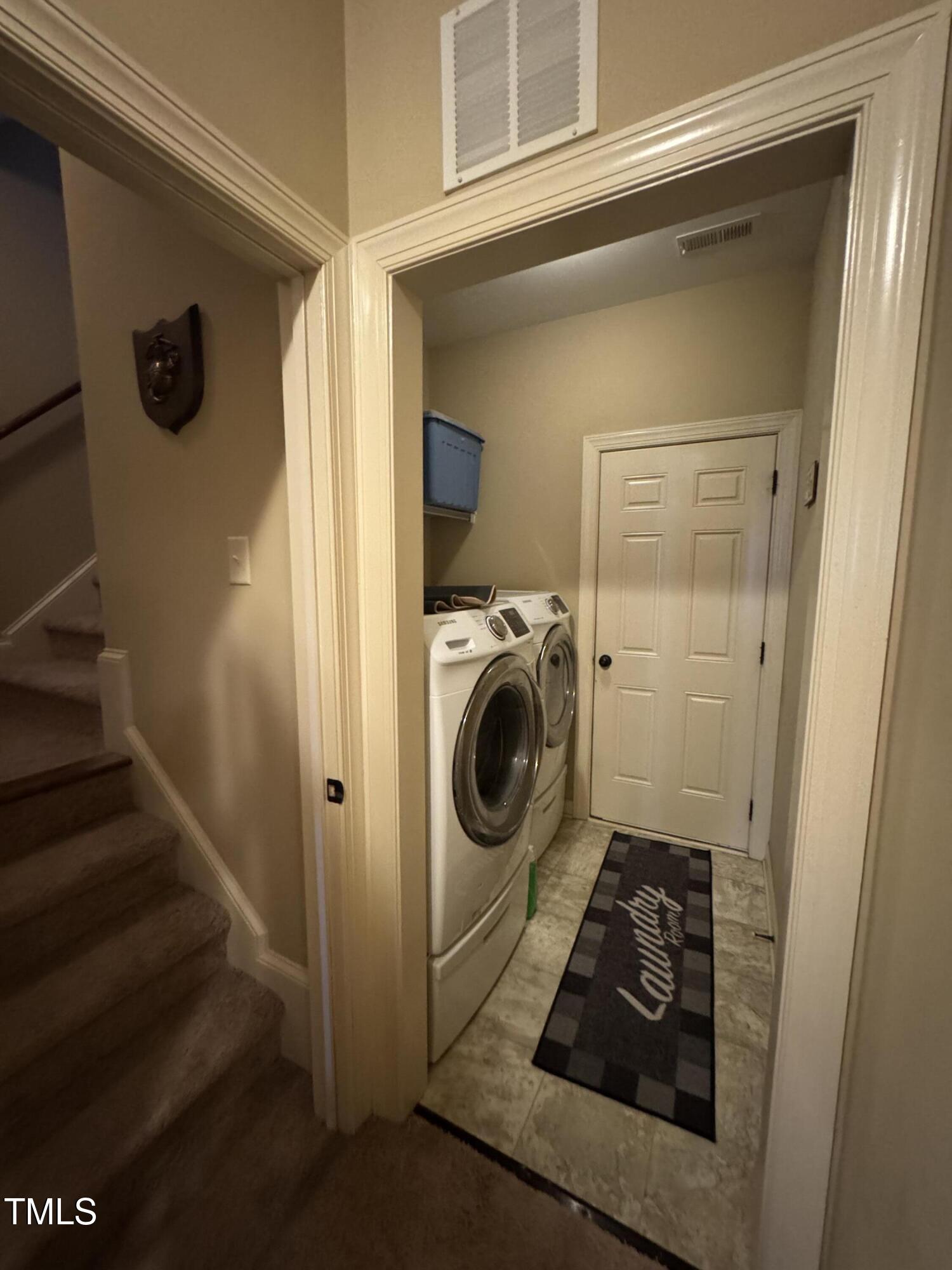 Laundry room with front-loading washer and dryer, tiled floor, and carpeted stairs nearby.