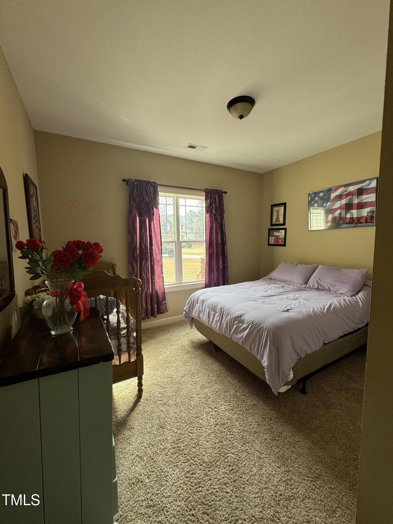 Bedroom with a lavender-covered bed, purple curtains, and red roses on a dark dresser.
