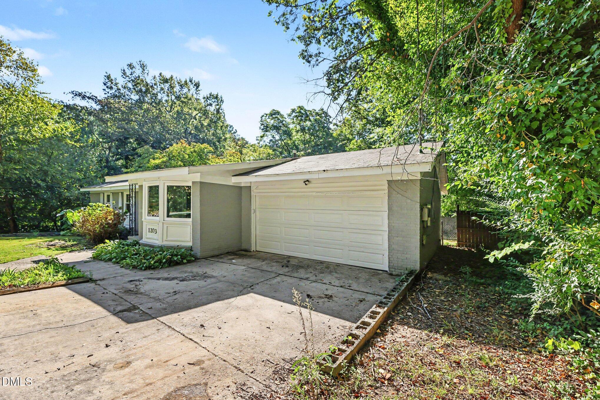 One-story light-colored home with an attached garage, concrete driveway, and mature trees.