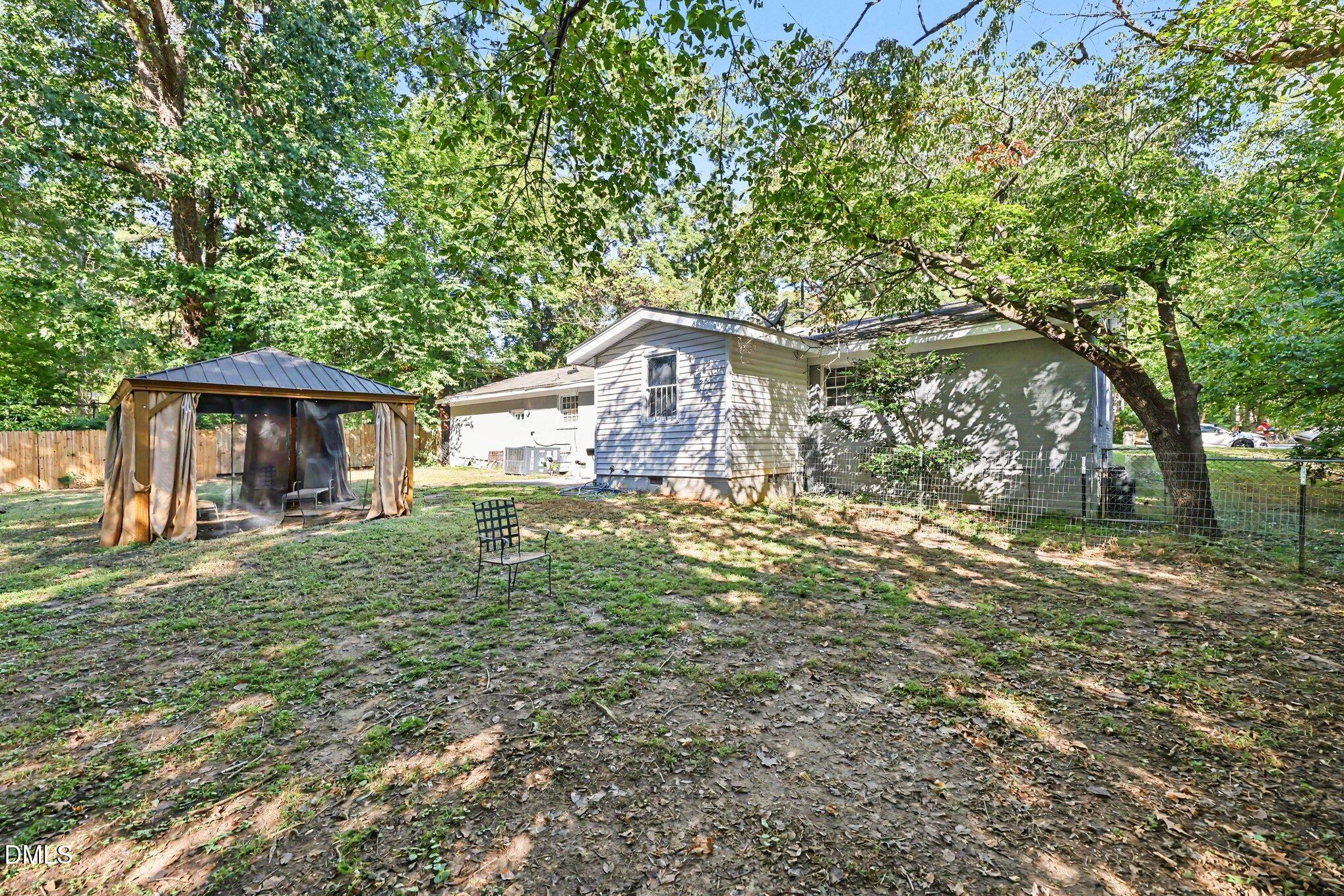 Rear of a gray house beneath mature trees, with a curtained gazebo in the yard.