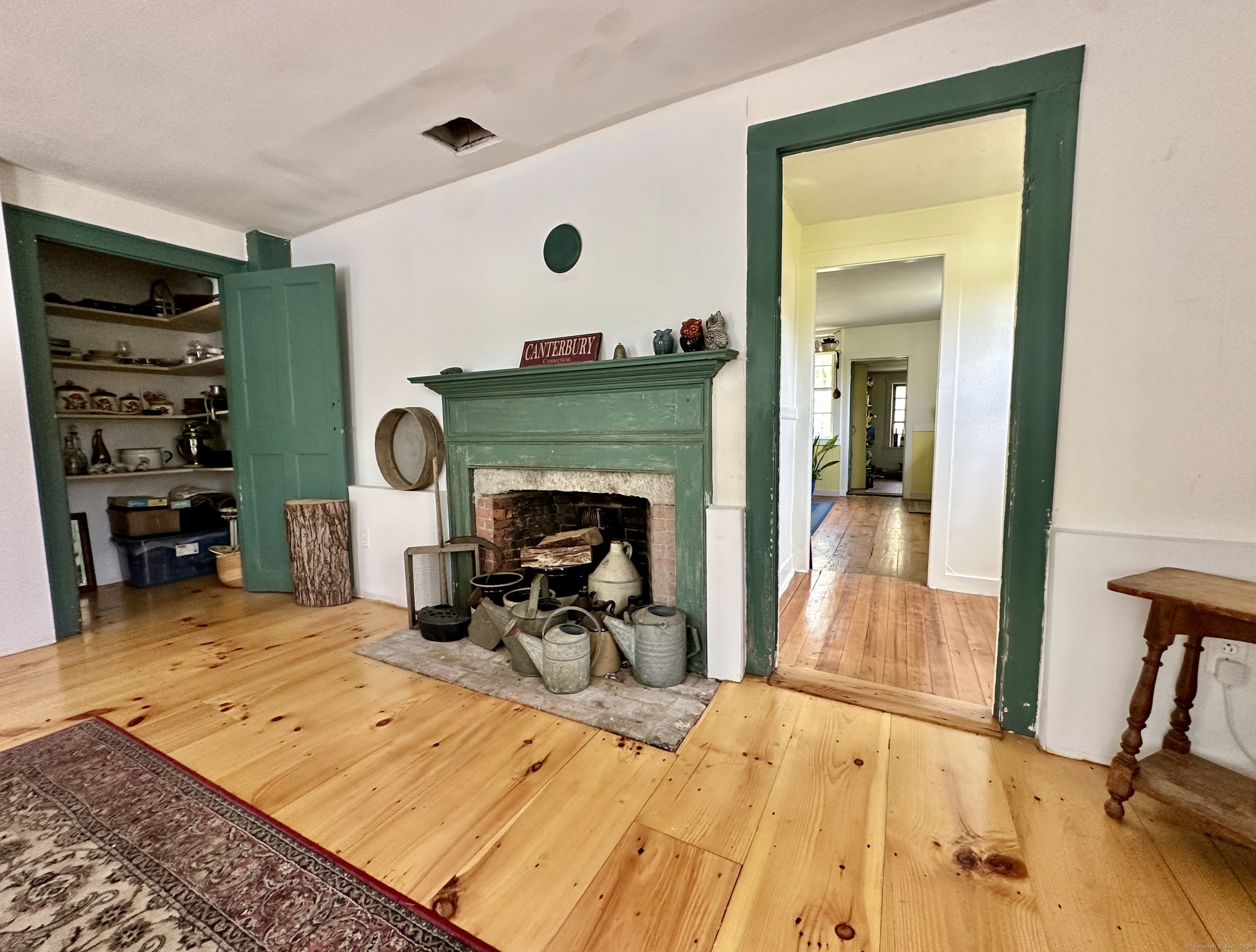 Room with a green-trimmed brick fireplace, wide-plank pine floors, and an open cupboard of pottery.
