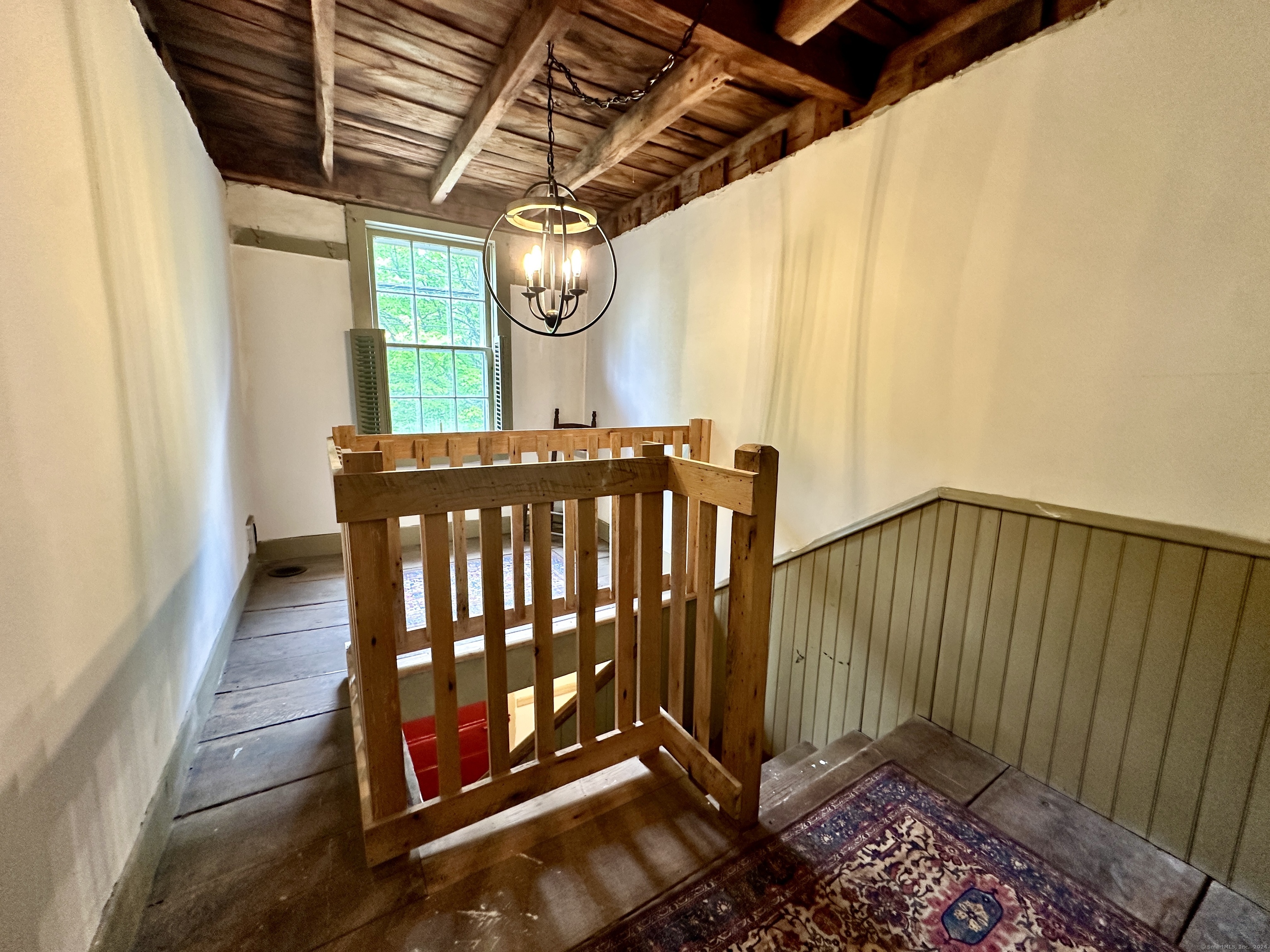 Stair landing with exposed timber beams, wooden railing, chandelier, and a tall multi-pane window.