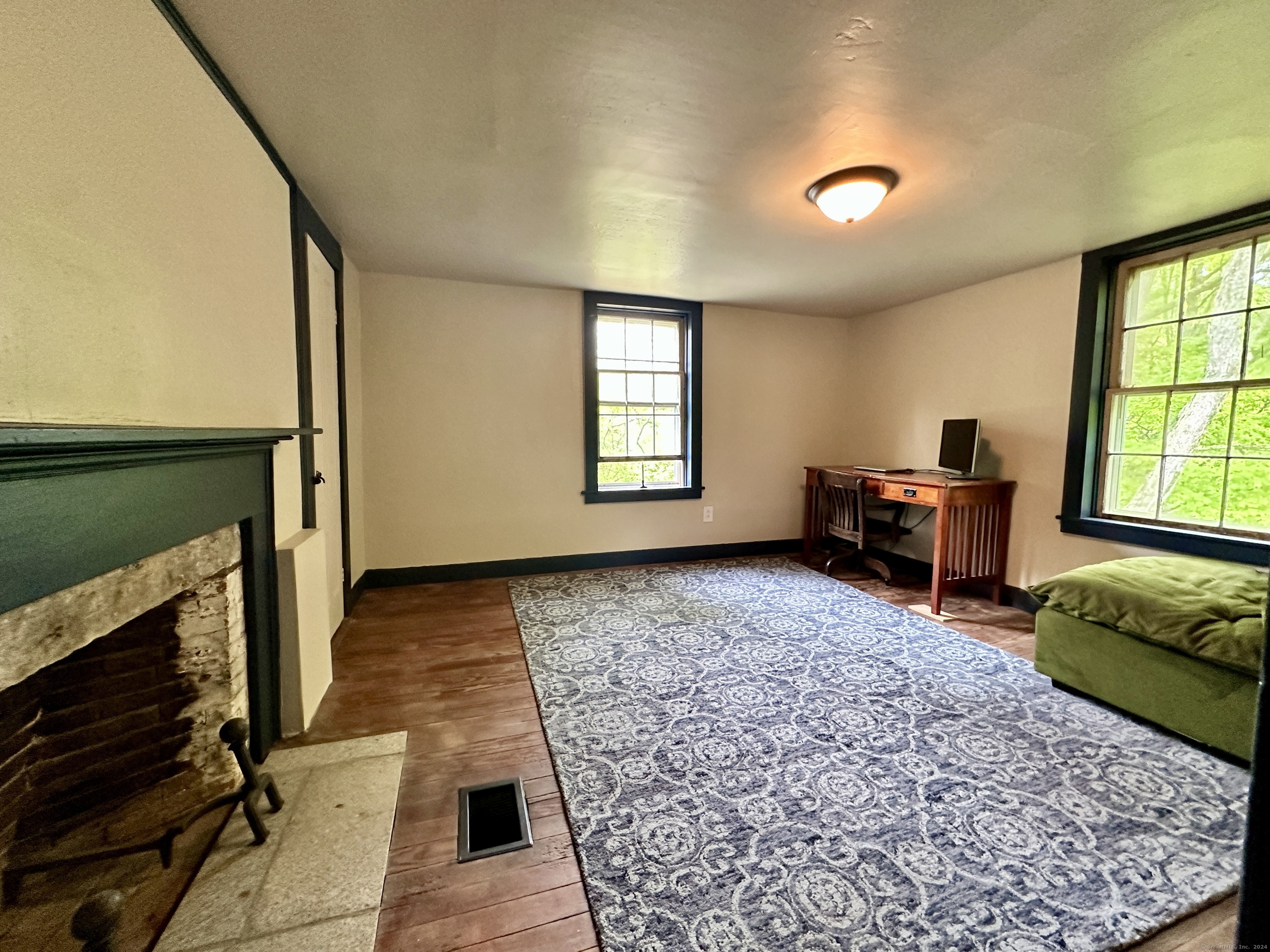 Room with a stone fireplace, hardwood floors, patterned rug, and desk beside leafy windows.