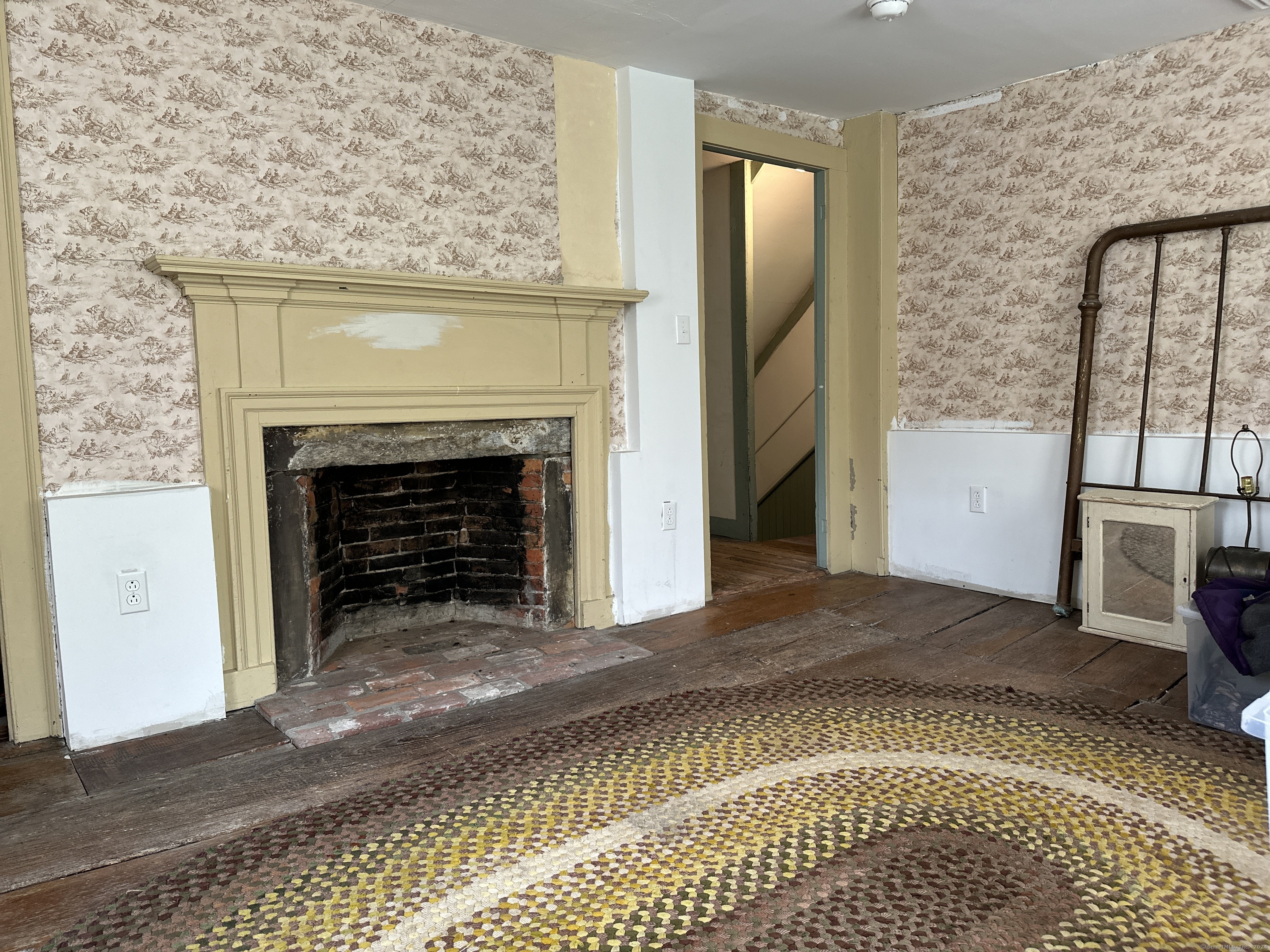 Room with a large beige fireplace, exposed brick hearth, patterned wallpaper, and worn plank floors.