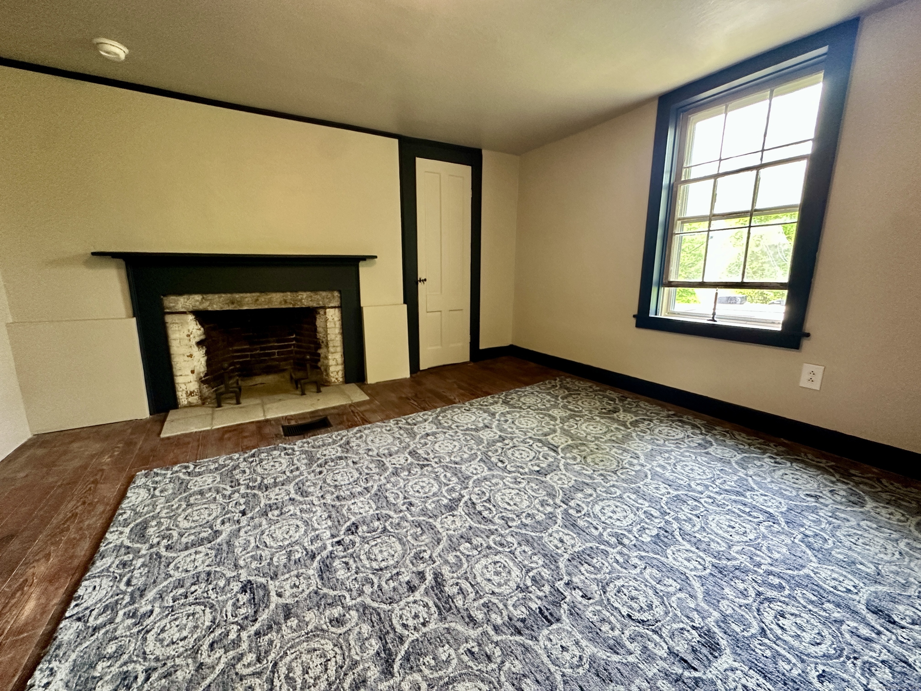 Unfurnished room with a dark fireplace, hardwood floors, patterned rug, and tall window.