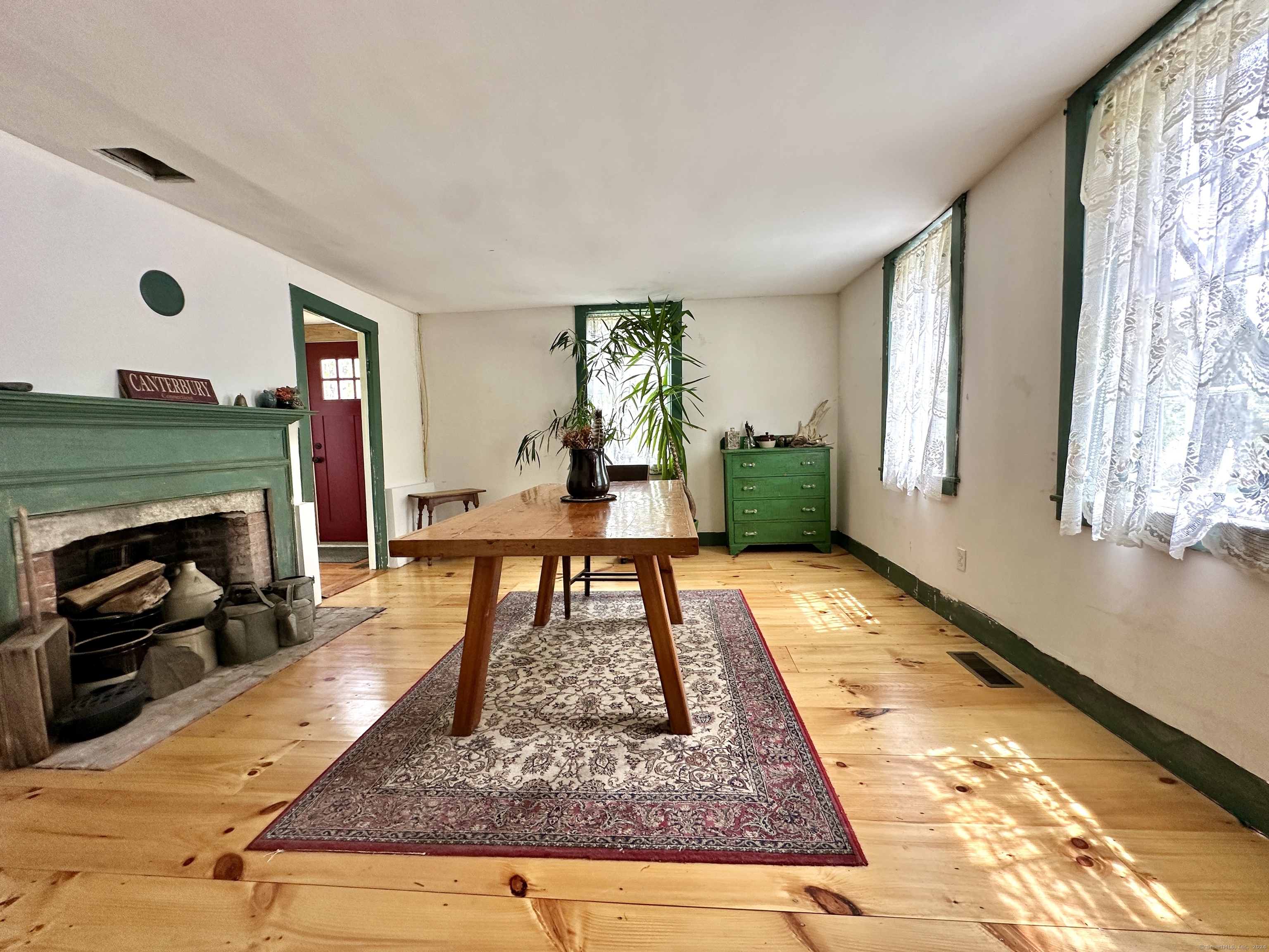 Dining room with a long wooden table, green fireplace, wide plank floors, and lace-curtained windows.