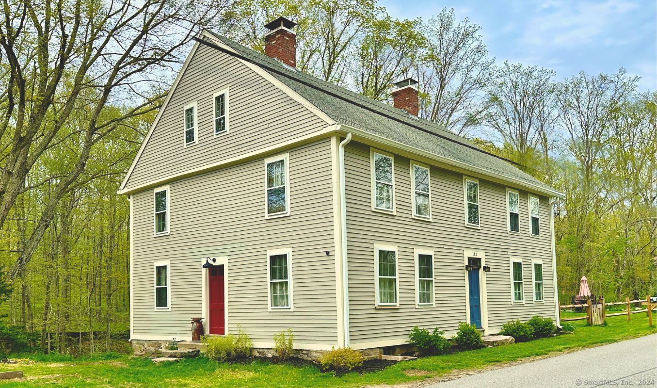 Two-story beige clapboard house with a steep roof, brick chimneys, and red and blue doors.
