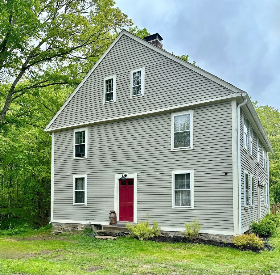 Gray two-story house with white trim and a red front door, beside a wooded area.