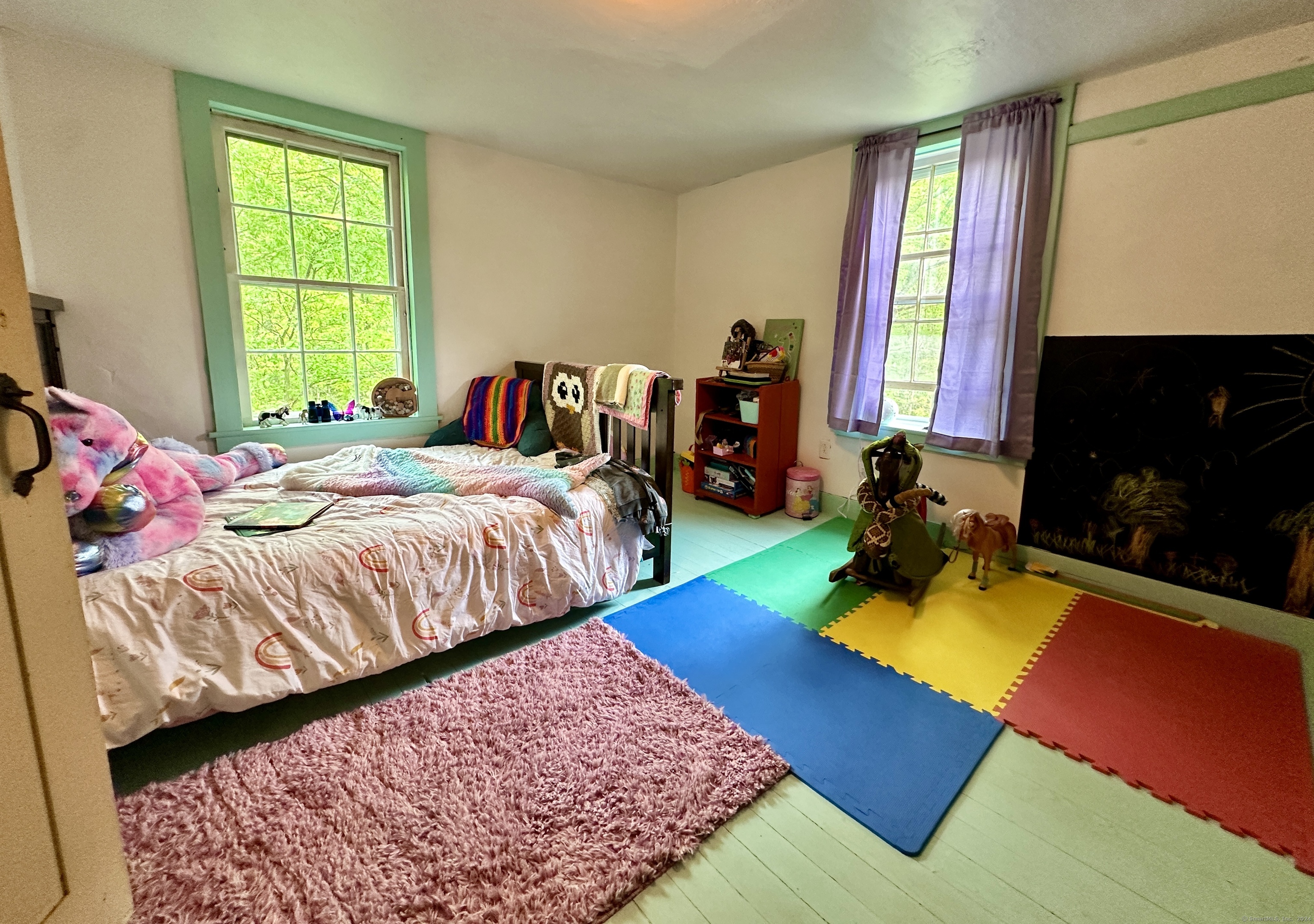 Bedroom with a bed, colorful play mats, toys, and two large green-trimmed windows.