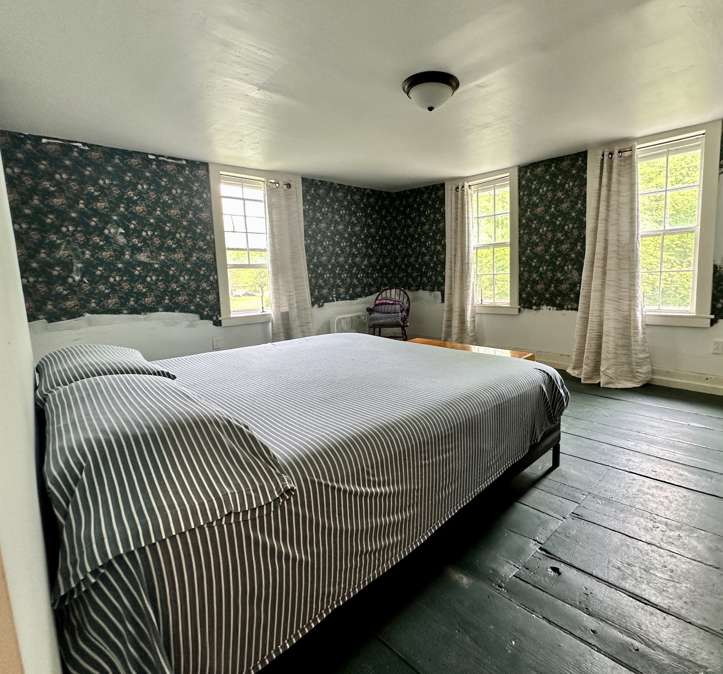 Bedroom with striped bedding, dark wood floors, floral wallpaper, and three bright windows.