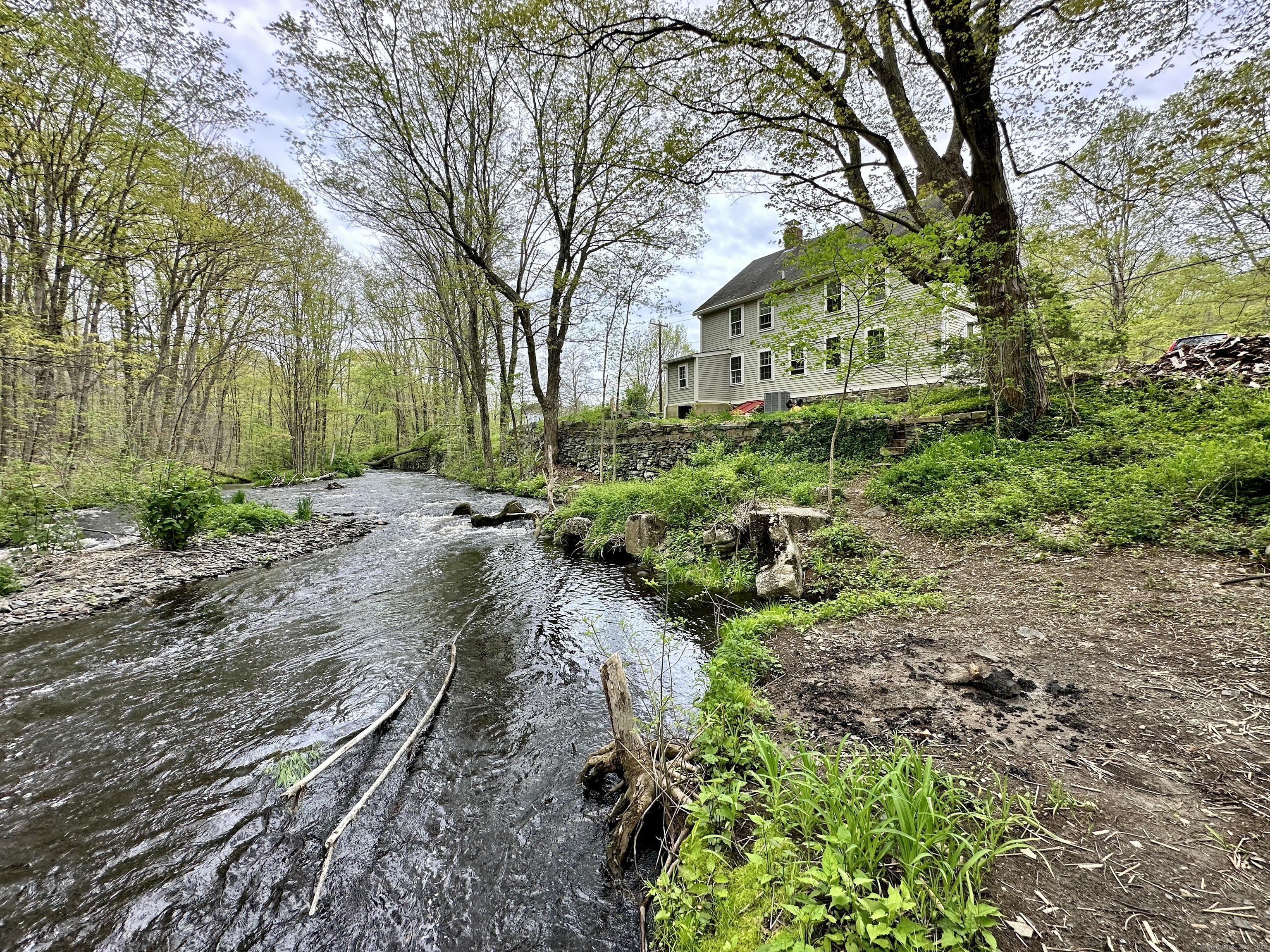 Two-story house above a wooded stream, with a stone retaining wall and dirt path along the bank.