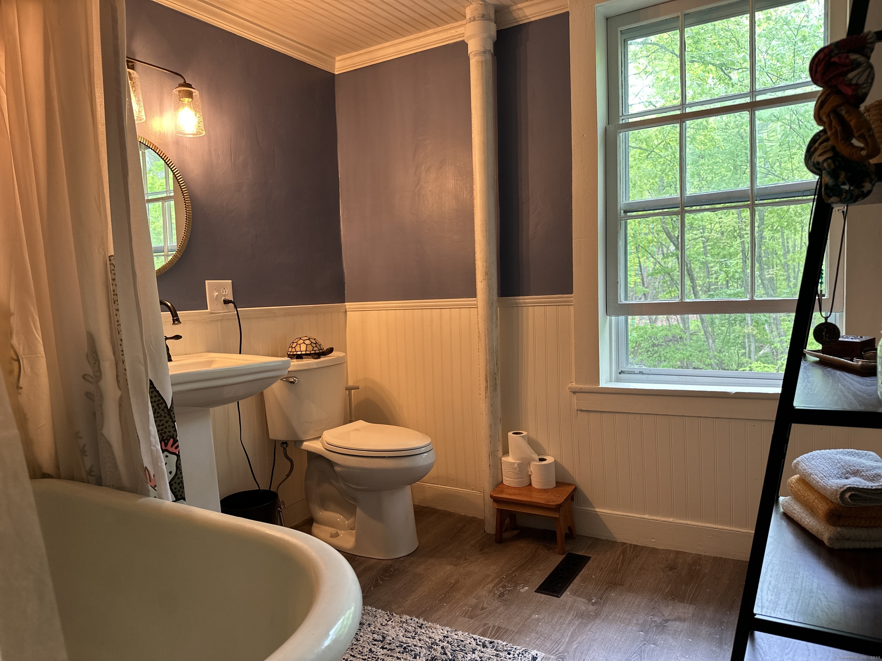 Bathroom with a freestanding tub, purple walls, white beadboard, and a large window overlooking trees.