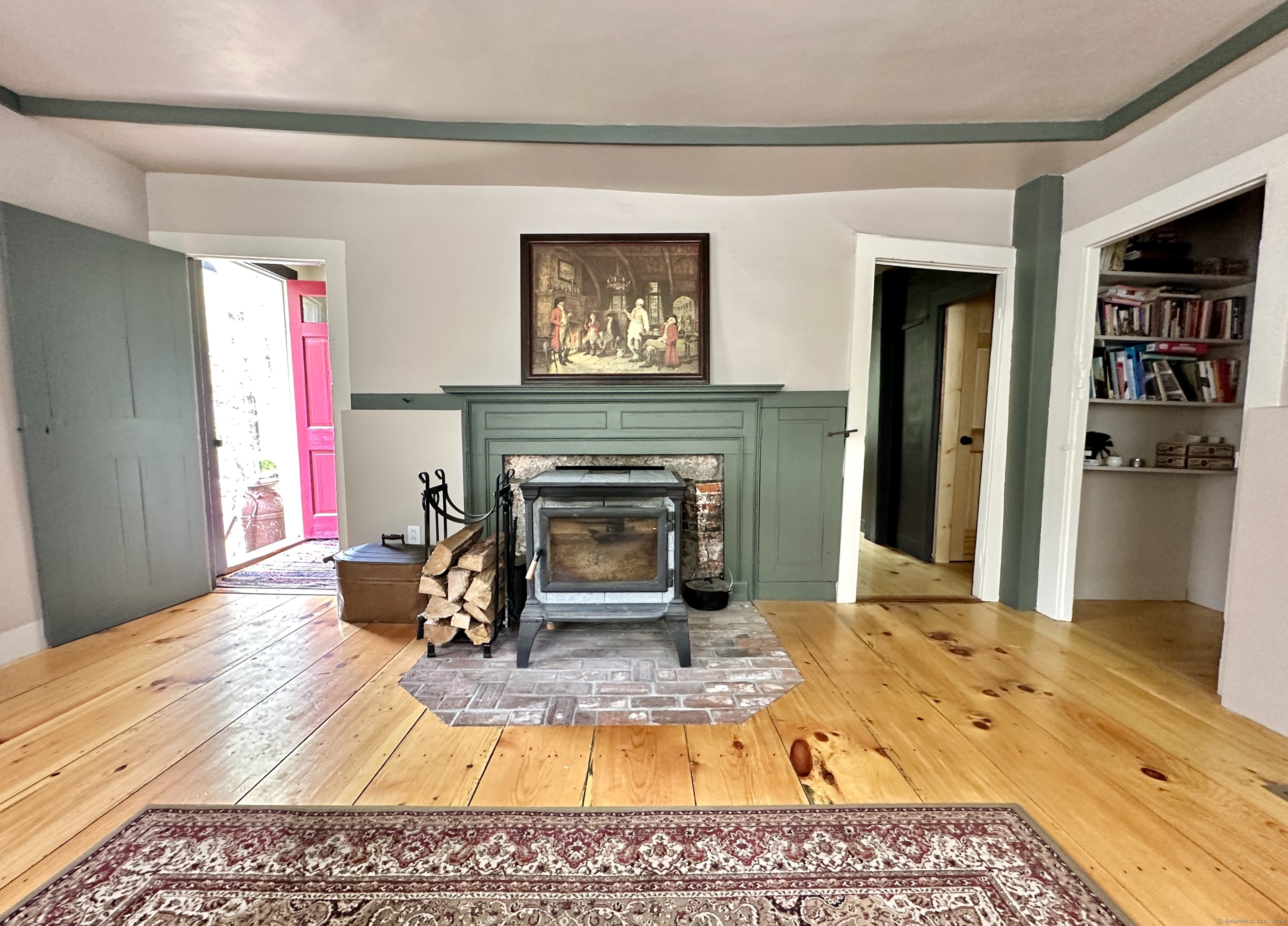Living room with a wood stove, green paneled fireplace, wide-plank floors, and an open pink door.