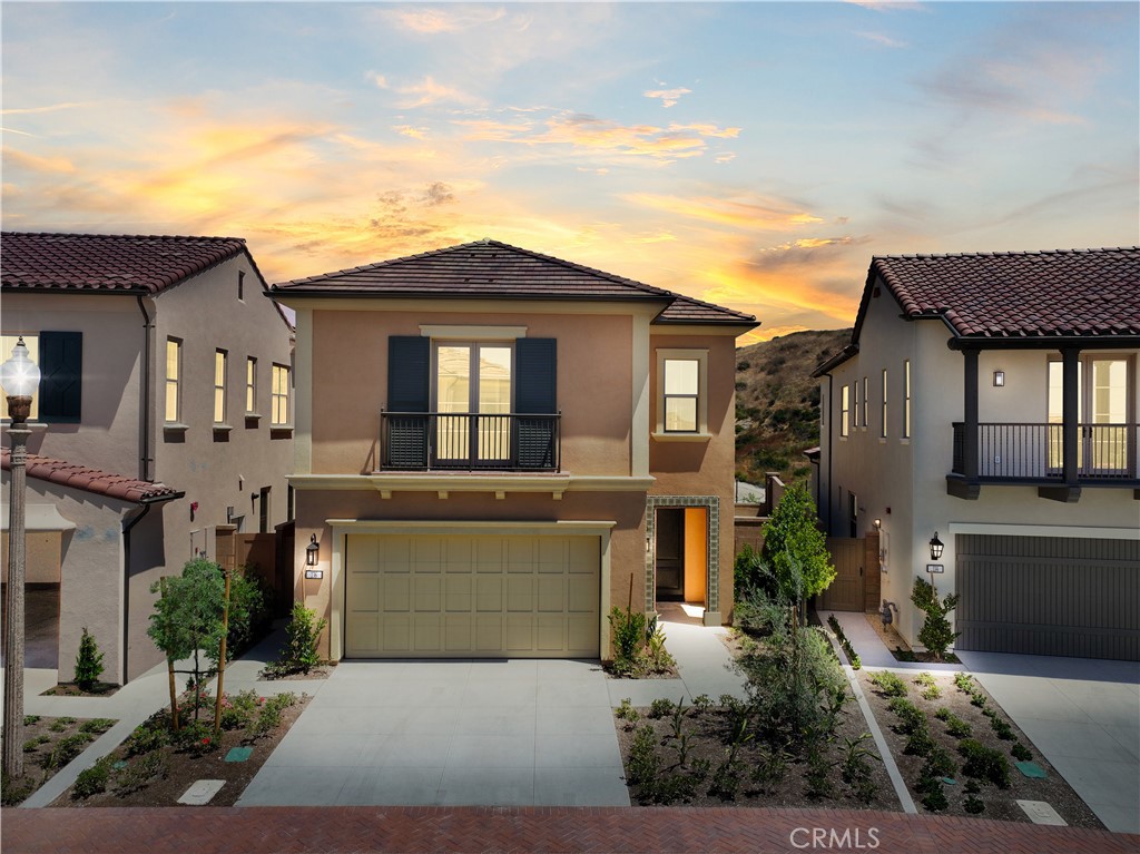 Two-story tan stucco home with a balcony and attached garage beneath a colorful sunset sky.