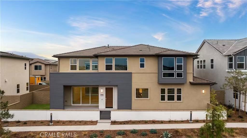 Two-story beige home with gray siding, large windows, and a low front patio wall.