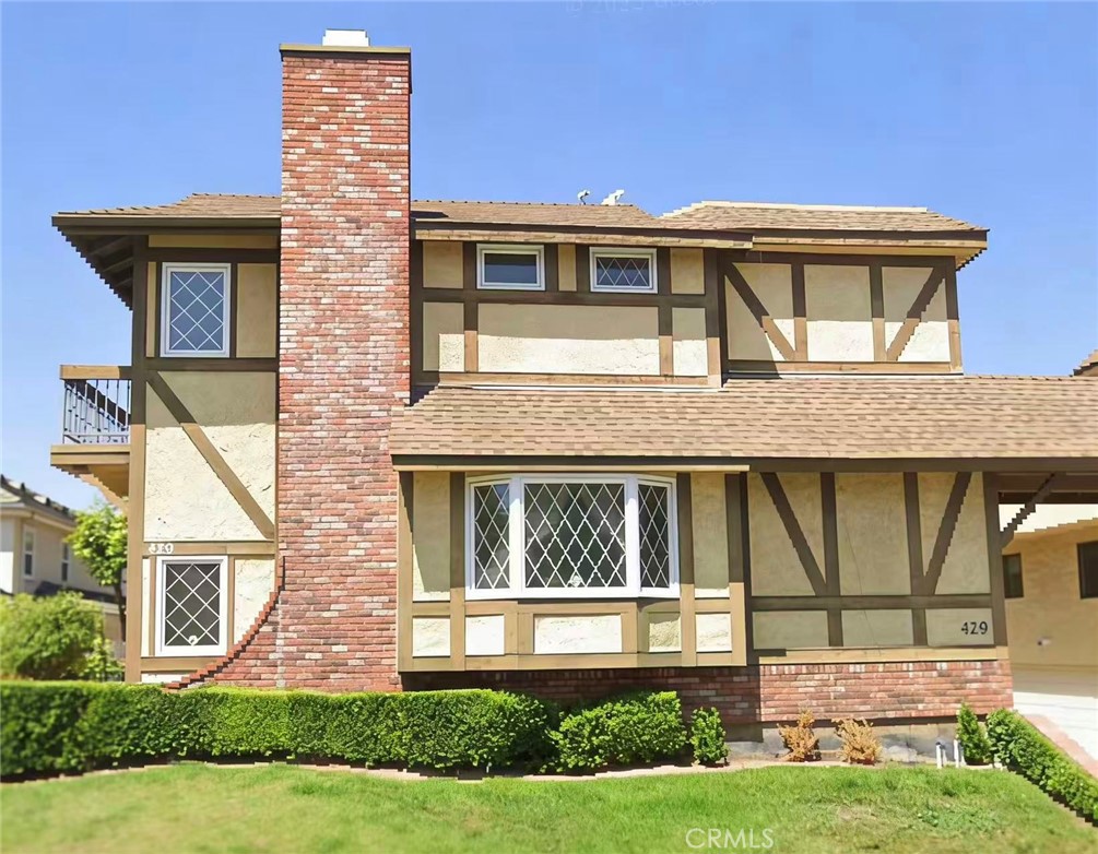 Two-story Tudor-style house with a tall brick chimney, diamond-pane windows, and trimmed front hedges.