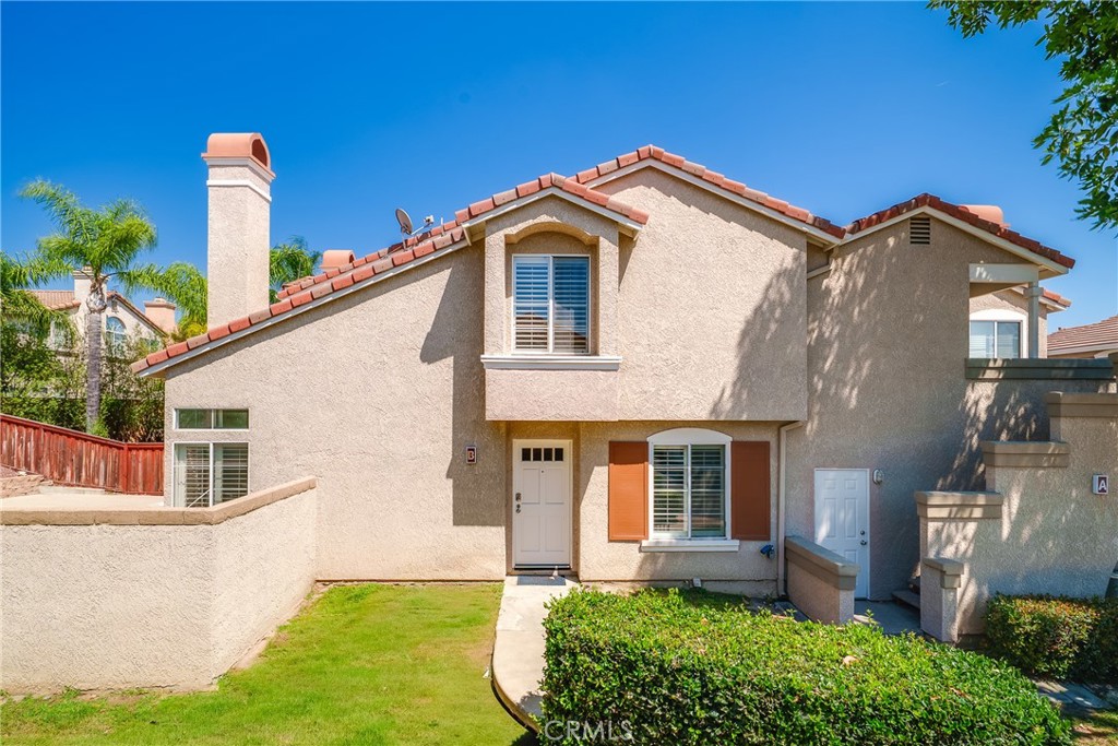 Two-story beige stucco house with a red tile roof, small front lawn, and trimmed shrubs.