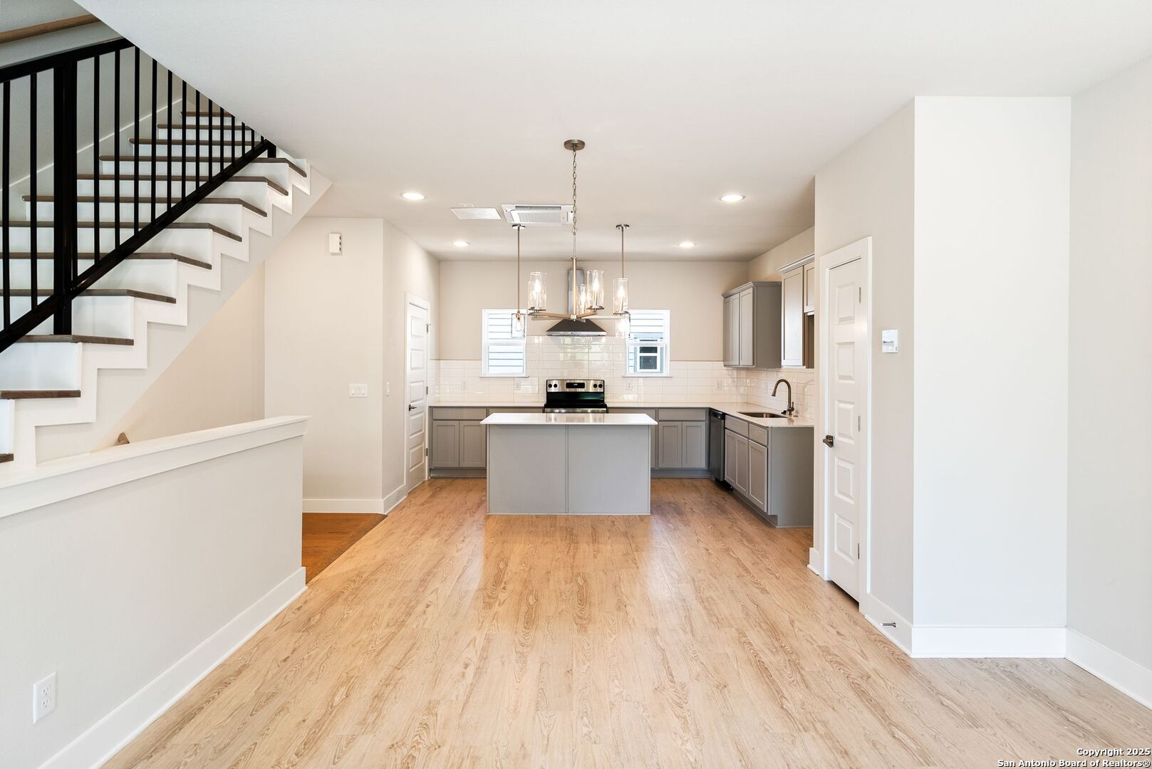 Open kitchen with a central island, gray cabinets, stainless appliances, and a dark-railed staircase.