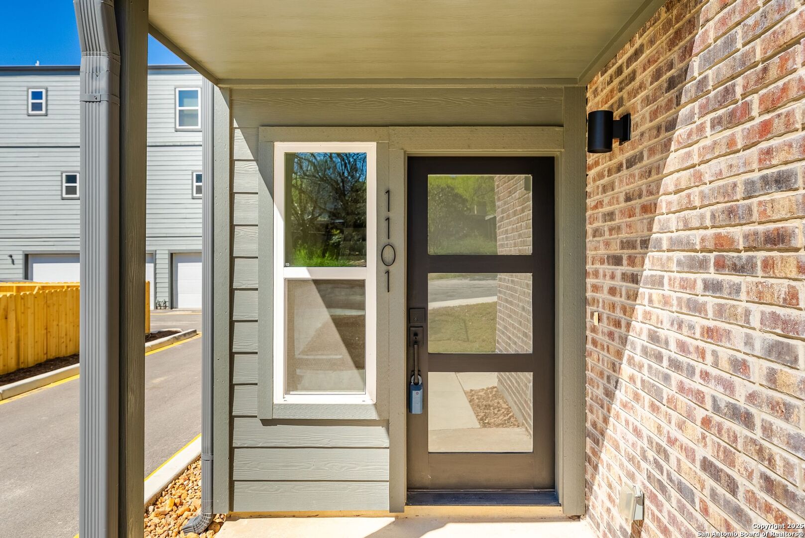 Covered front entry with a dark glass-paneled door, sidelight, gray-green siding, and brick wall.