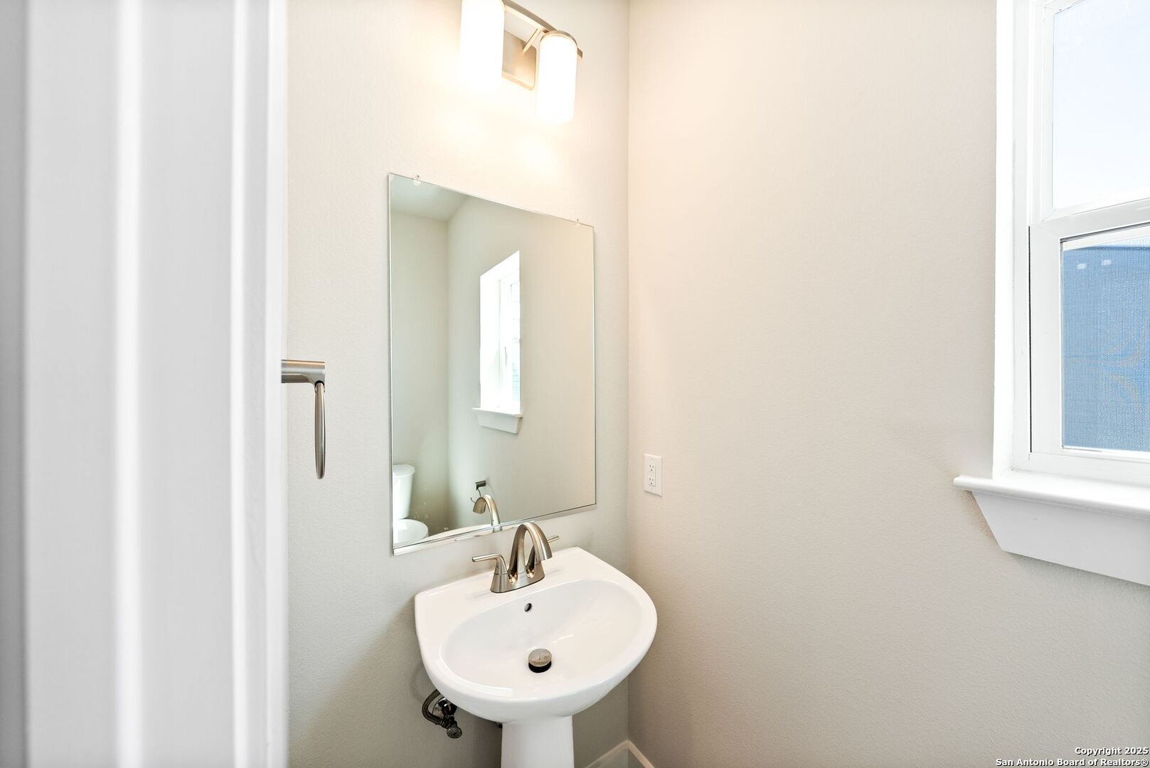 Bathroom pedestal sink with brushed metal faucet beneath a wall mirror, beside a window.