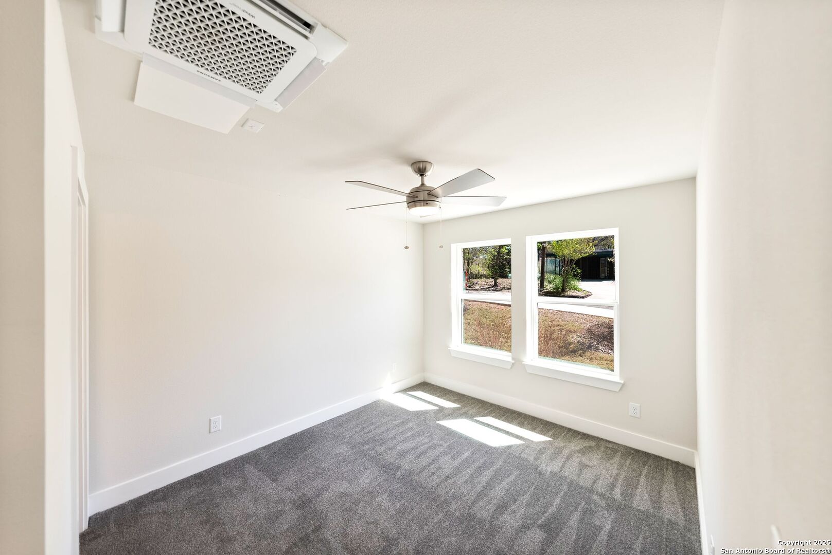 Empty bedroom with gray carpet, a ceiling fan, and two windows overlooking the yard.