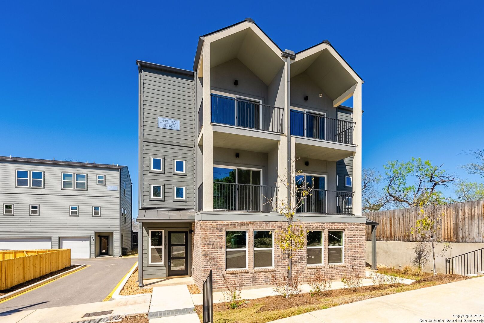 Modern multi-story condominium building with stacked balconies, gray siding, and a brick ground floor.