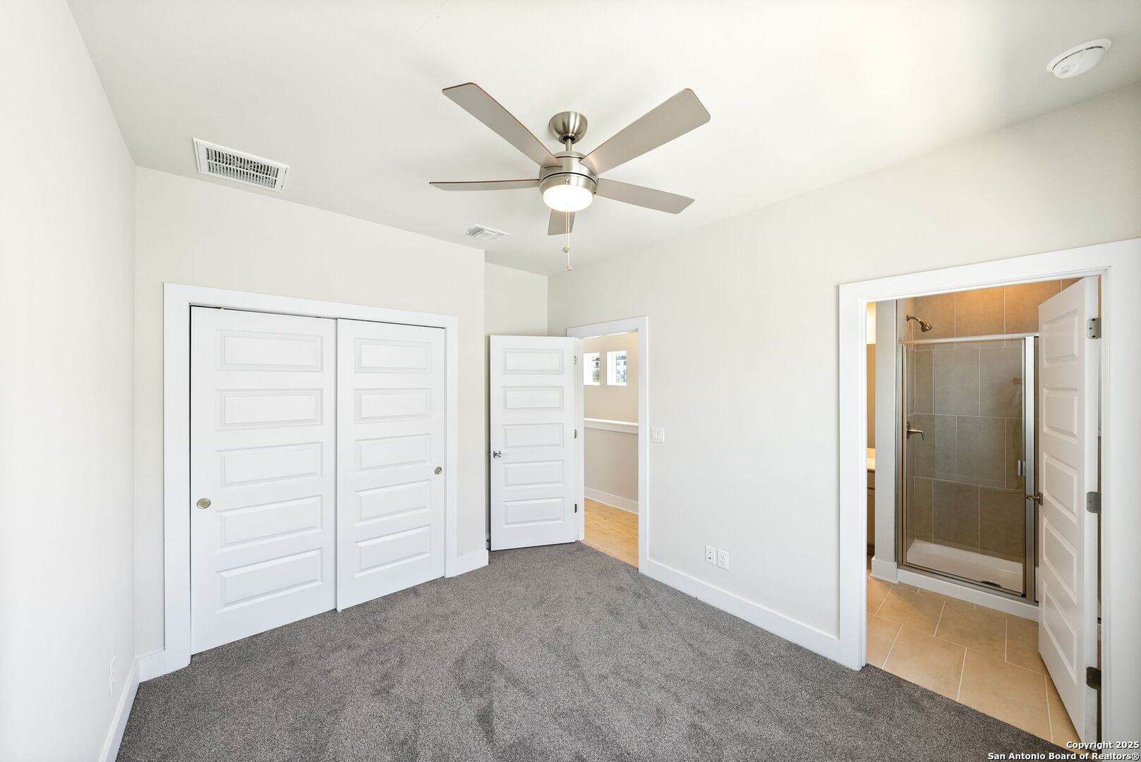 Unfurnished bedroom with gray carpet, ceiling fan, double closets, and an adjoining bathroom with a glass shower.