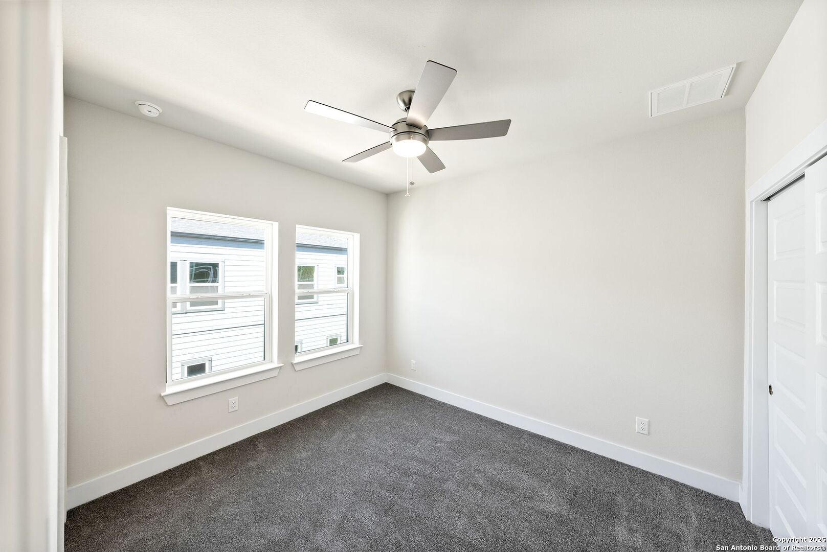 Empty bedroom with gray carpet, two windows, and a ceiling fan with a light.