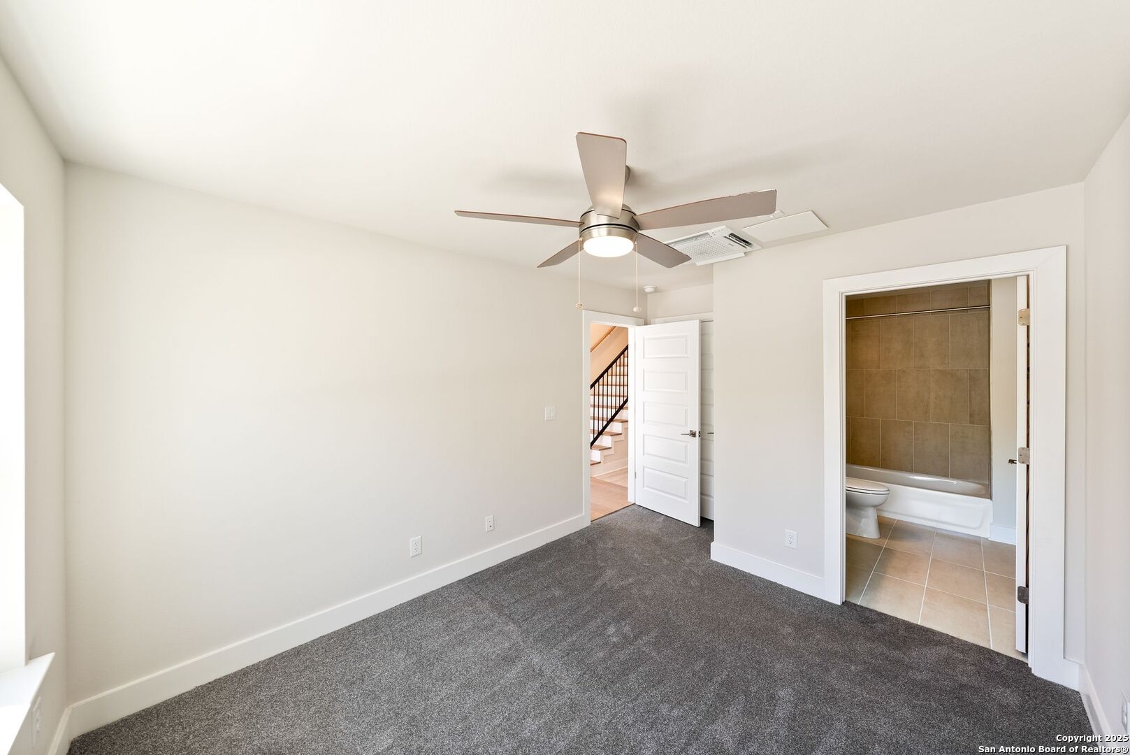 Empty bedroom with gray carpet and ceiling fan, opening to a tiled bathroom and staircase.
